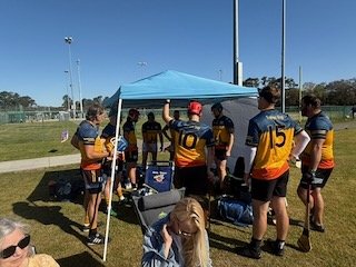 A youth football team gathered under a blue canopy tent on a sports field, wearing matching jerseys and shorts, with some players standing and others sitting nearby, alongside a dog and sports equipment.
