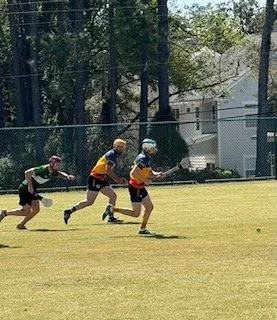 Four children playing tag on a grassy field, with a chain-link fence and trees in the background.