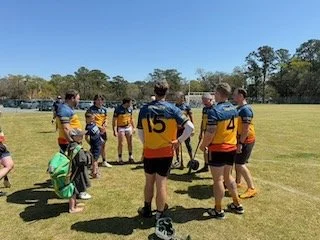 Group of young hurling players gathered on a field during daytime practice or game, with trees in the background.