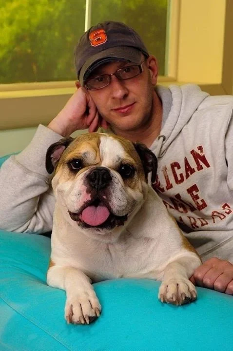 A man with glasses and a cap sitting behind a happy bulldog on a teal cushion in a room with green trees visible through the window.