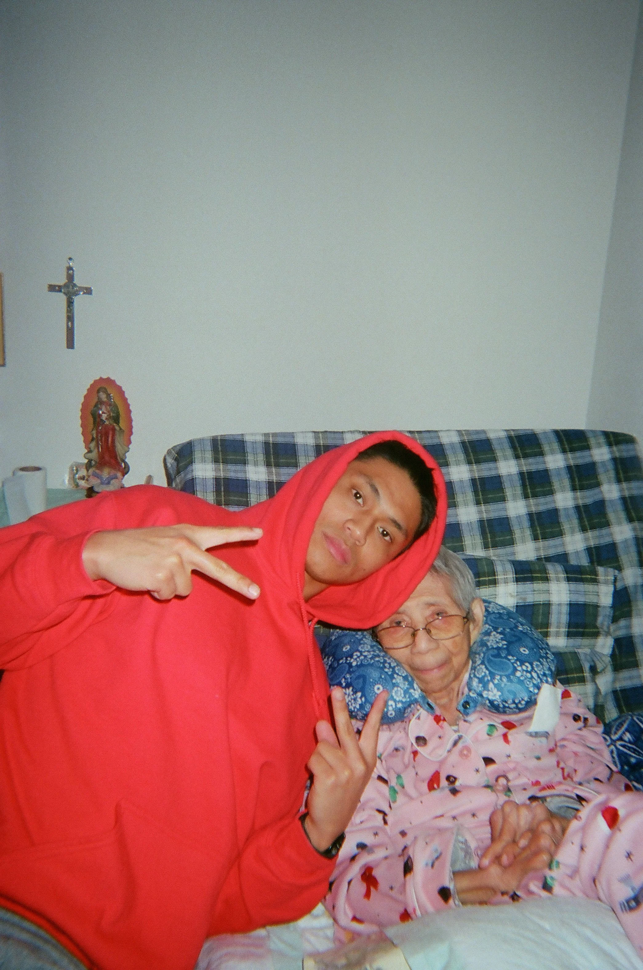 Chris Punsalan wearing a red hoodie and posing beside his grandmother as she rests in bed at home.