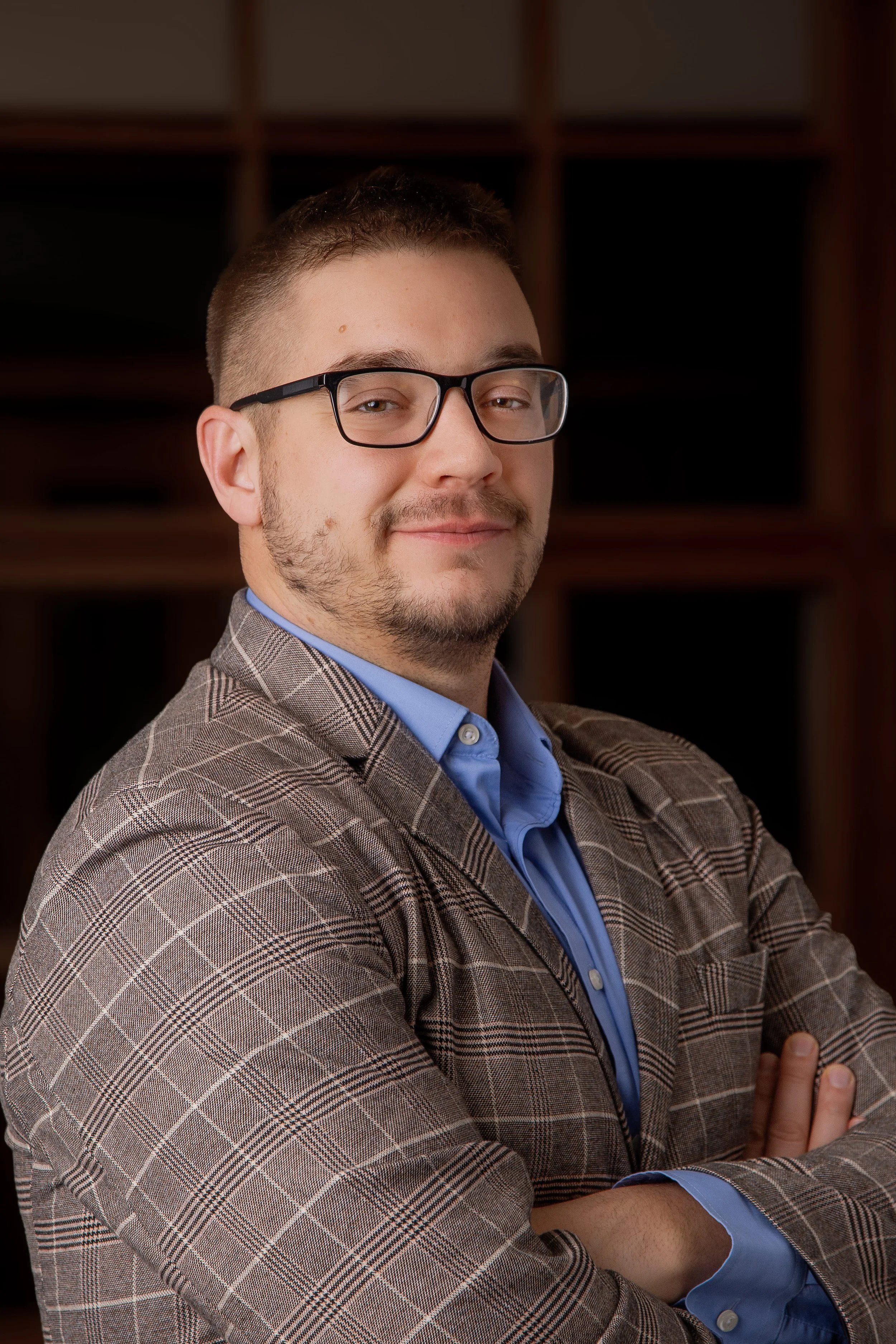 Portrait of a young man with short hair and glasses, wearing a checked blazer and blue shirt, standing with arms crossed in front of a dark background.