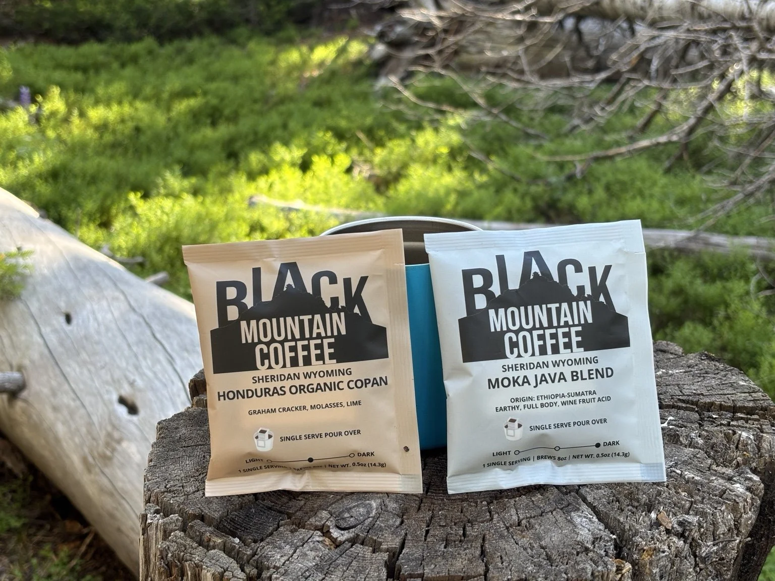 Two packets of Black Mountain Coffee placed on a tree stump outdoors with a background of lush green grass and tree branches.