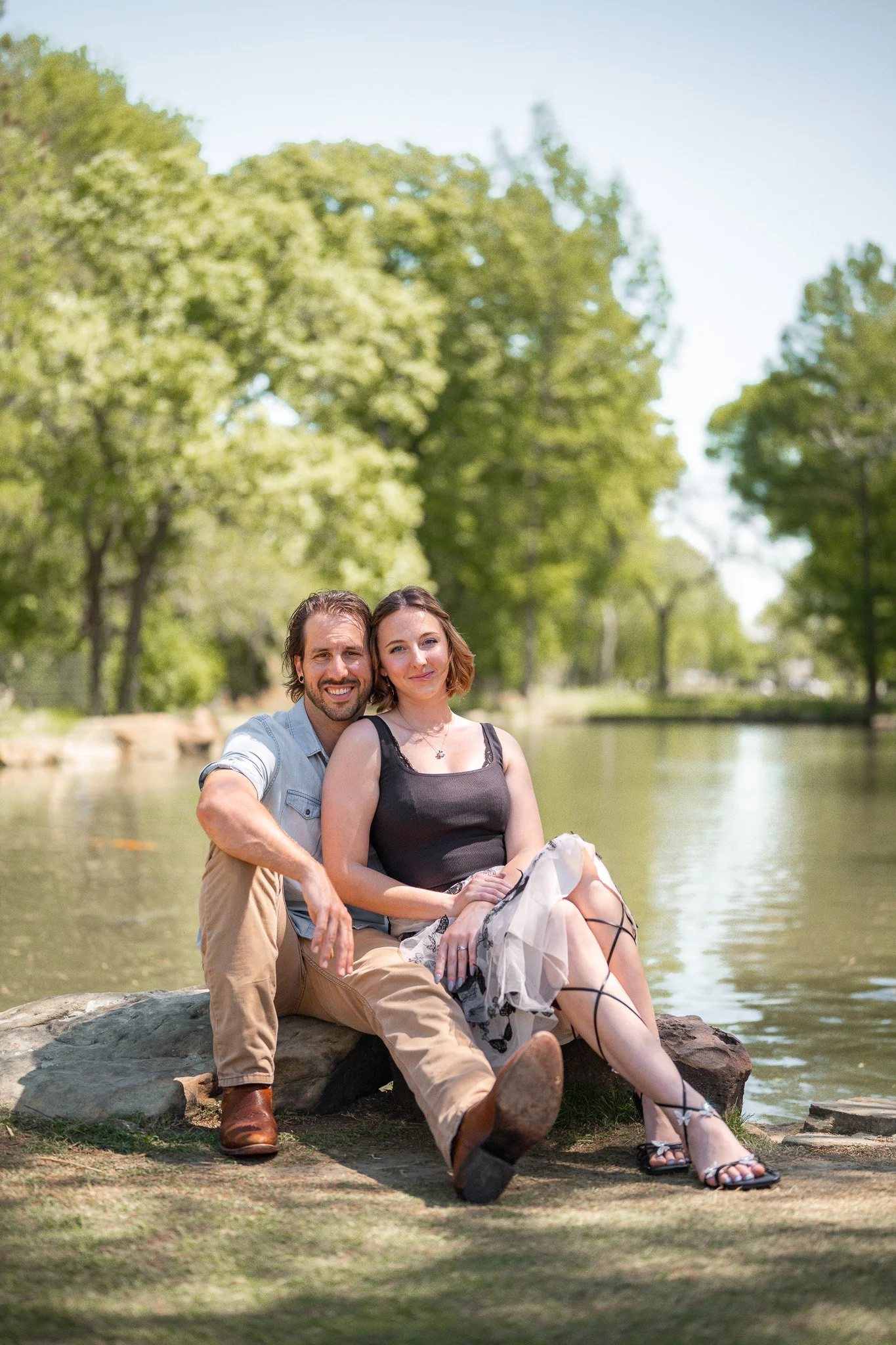 Couple sitting together on a rock beside a tree-lined pond in a park