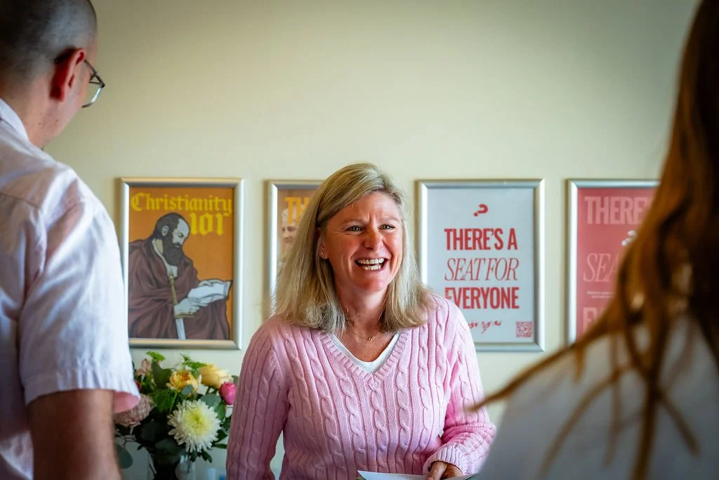 A woman in a pink jumper sharing conversation at St Philips, with an Alpha welcome poster behind her.