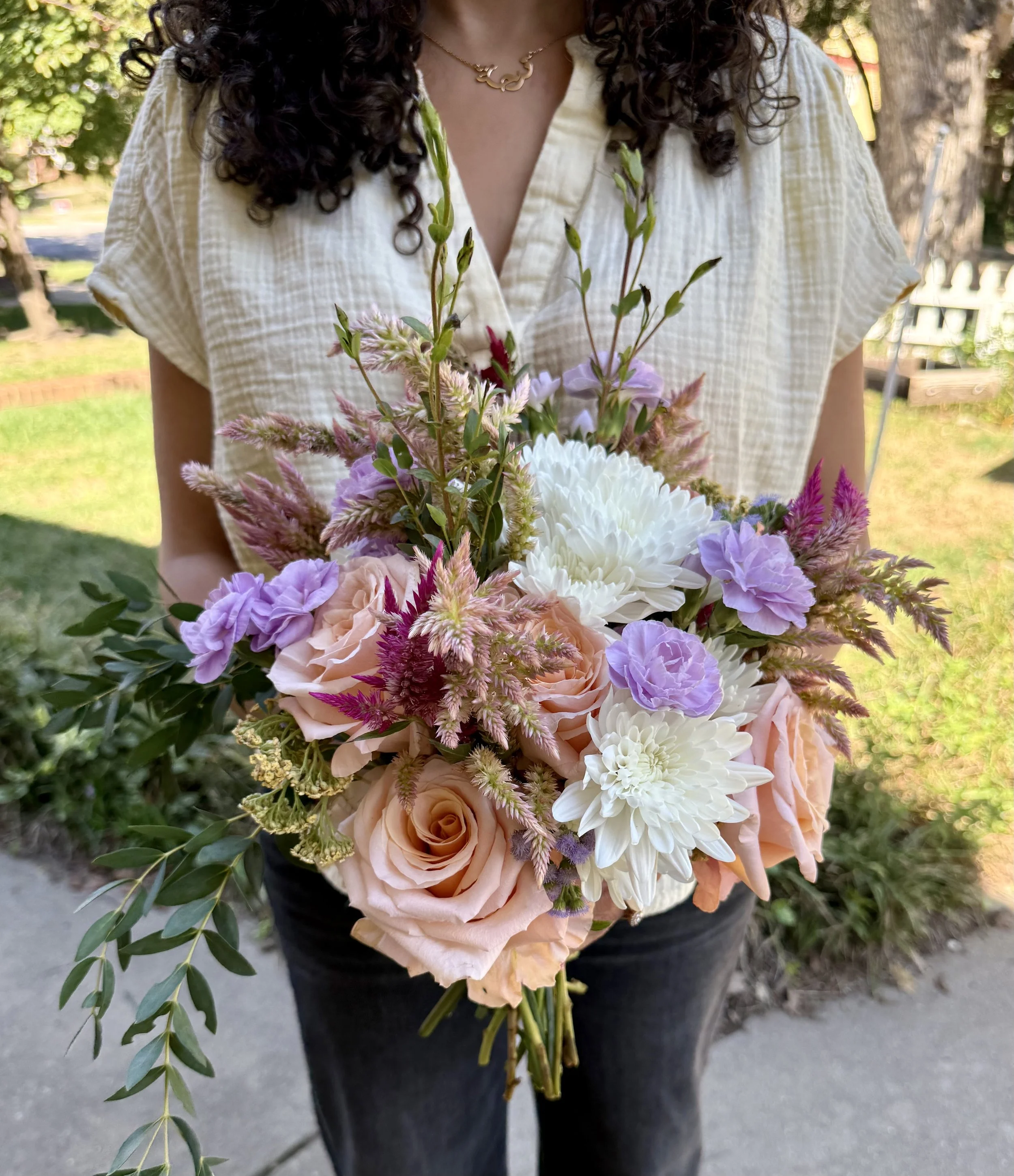 Person holding a large bouquet of colorful flowers outdoors, with trees and a grassy area in the background.