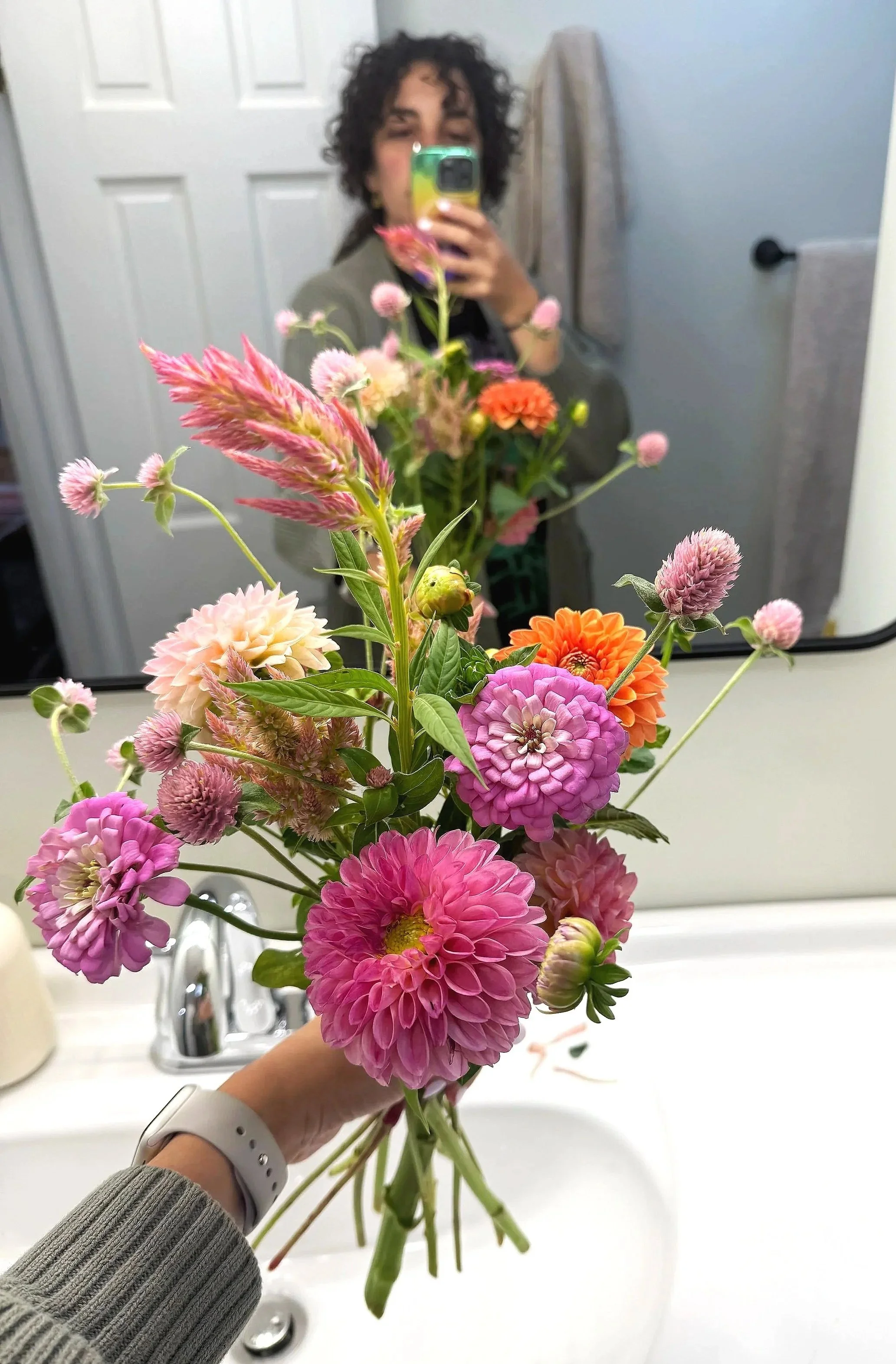A person with curly dark hair, wearing a gray sweater and a white smartwatch, holding a bouquet of pink, orange, purple, and white flowers in a bathroom mirror.