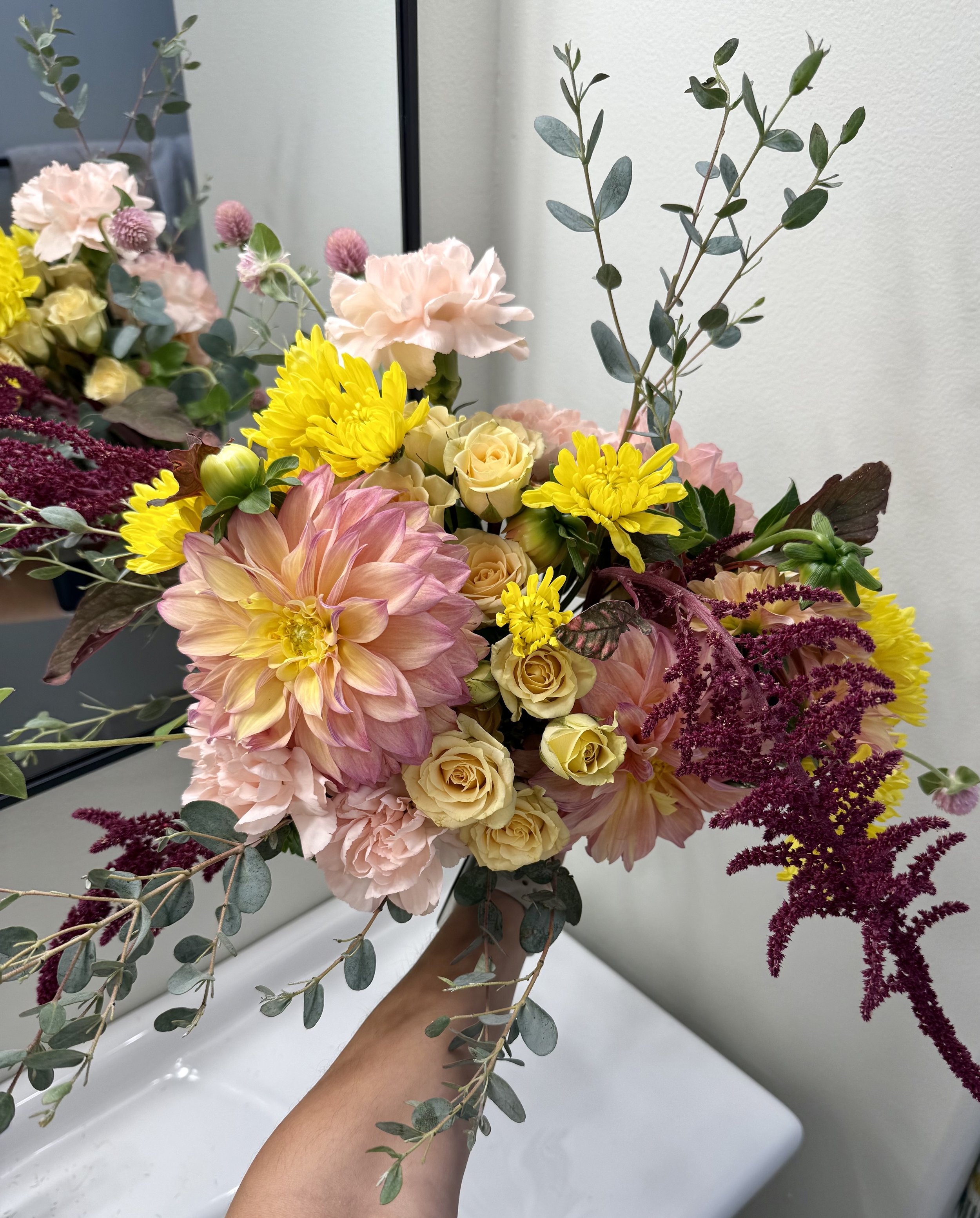 A bouquet of flowers held by a person's hand. The bouquet includes pale pink carnations, yellow chrysanthemums, roses, pink dahlias, purple amaranthus, and eucalyptus leaves, set against a plain white background.