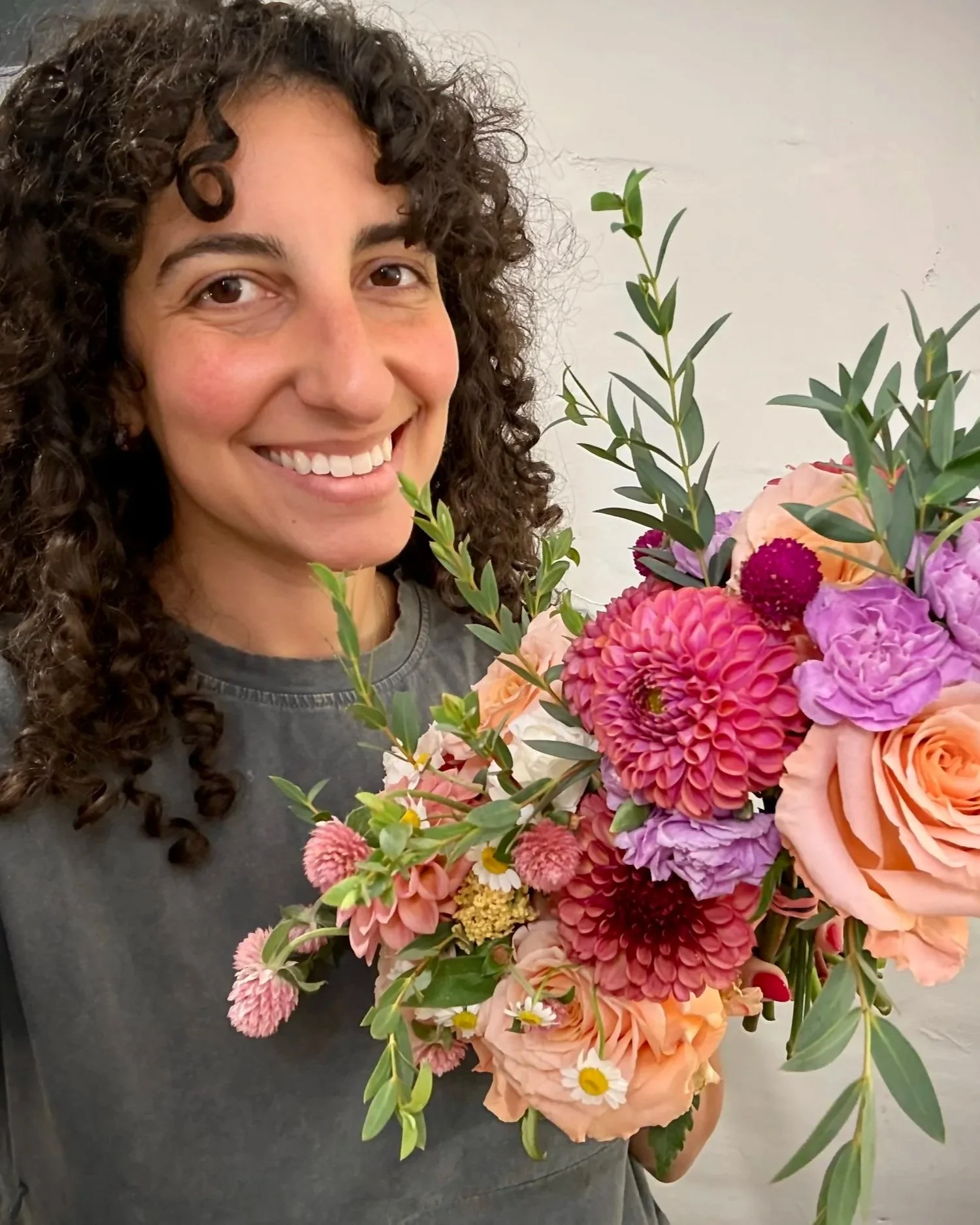 A woman with curly brown hair holding a large colorful bouquet of mixed flowers including pink, peach, purple, and white blooms, smiling at the camera.