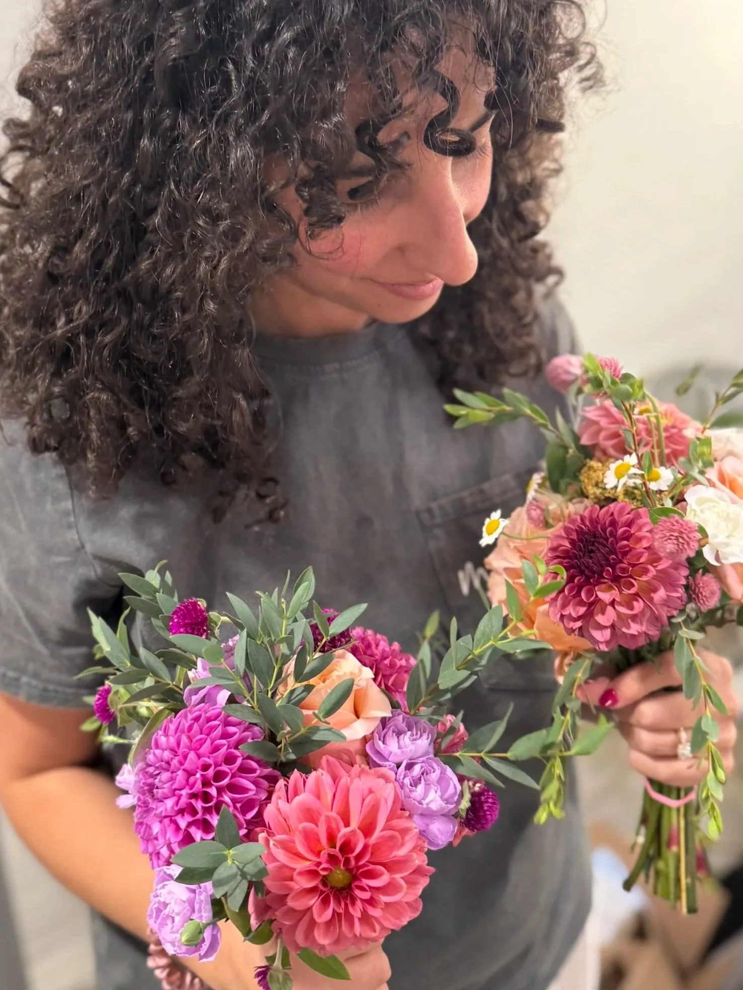 A woman with curly dark hair holds a colorful bouquet of pink, purple, and orange flowers with green leaves.