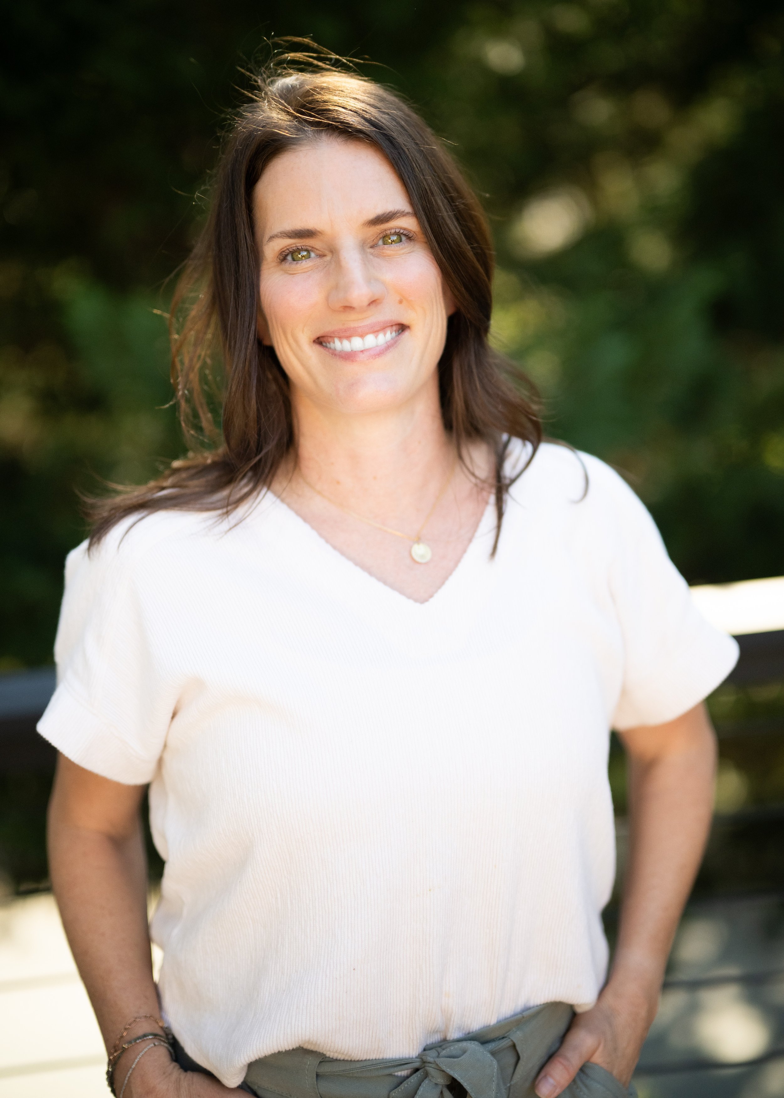 A woman with shoulder-length brown hair, light green eyes, and a bright smile, wearing a white V-neck shirt and gray pants, standing outdoors with a blurred green leafy background.