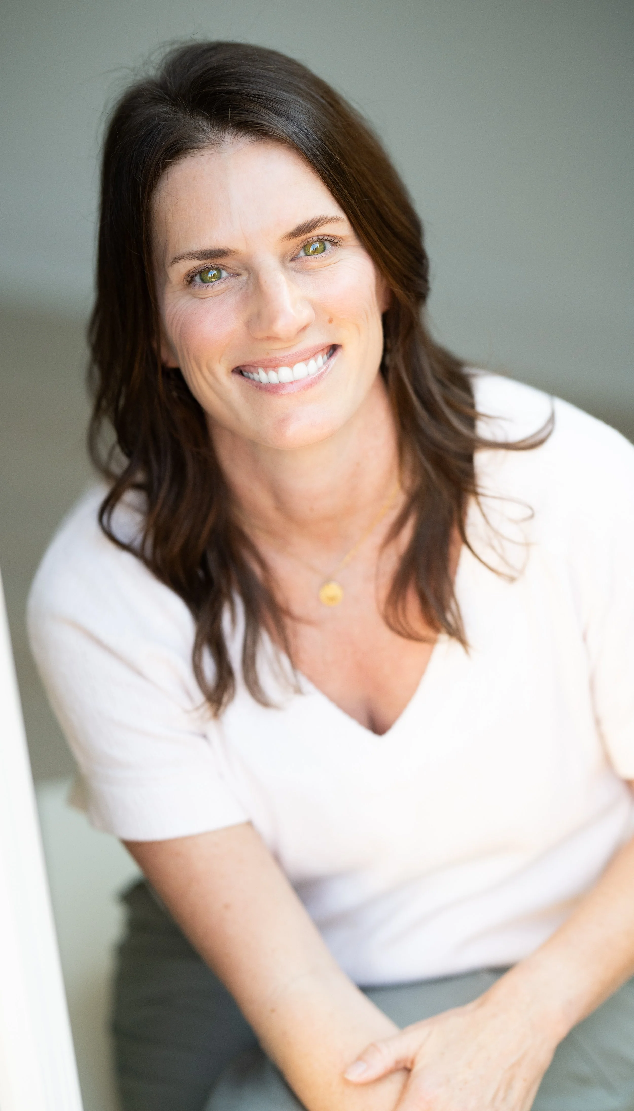 Close-up of a woman with shoulder-length brown hair, green eyes, and a bright smile, wearing a white top and a gold necklace, sitting indoors with a neutral gray background.