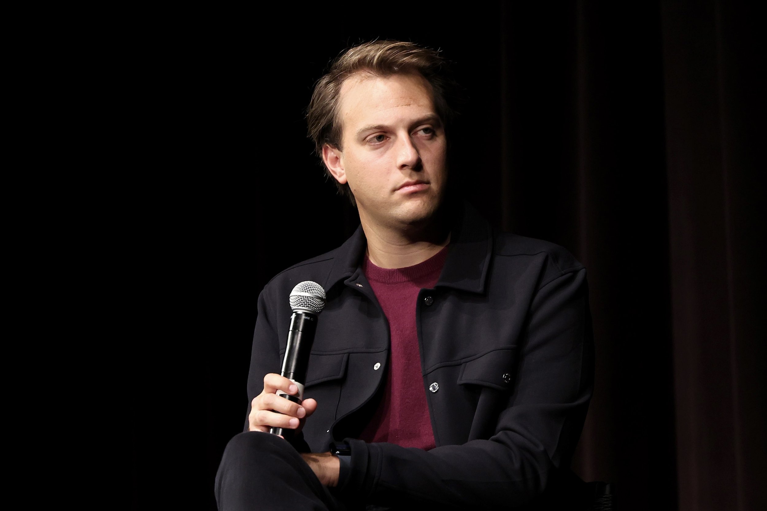 A man with light skin and brown hair is sitting, holding a microphone in his right hand. He is wearing a dark jacket over a red shirt and is looking to his left. The background is dark with some vertical lines.