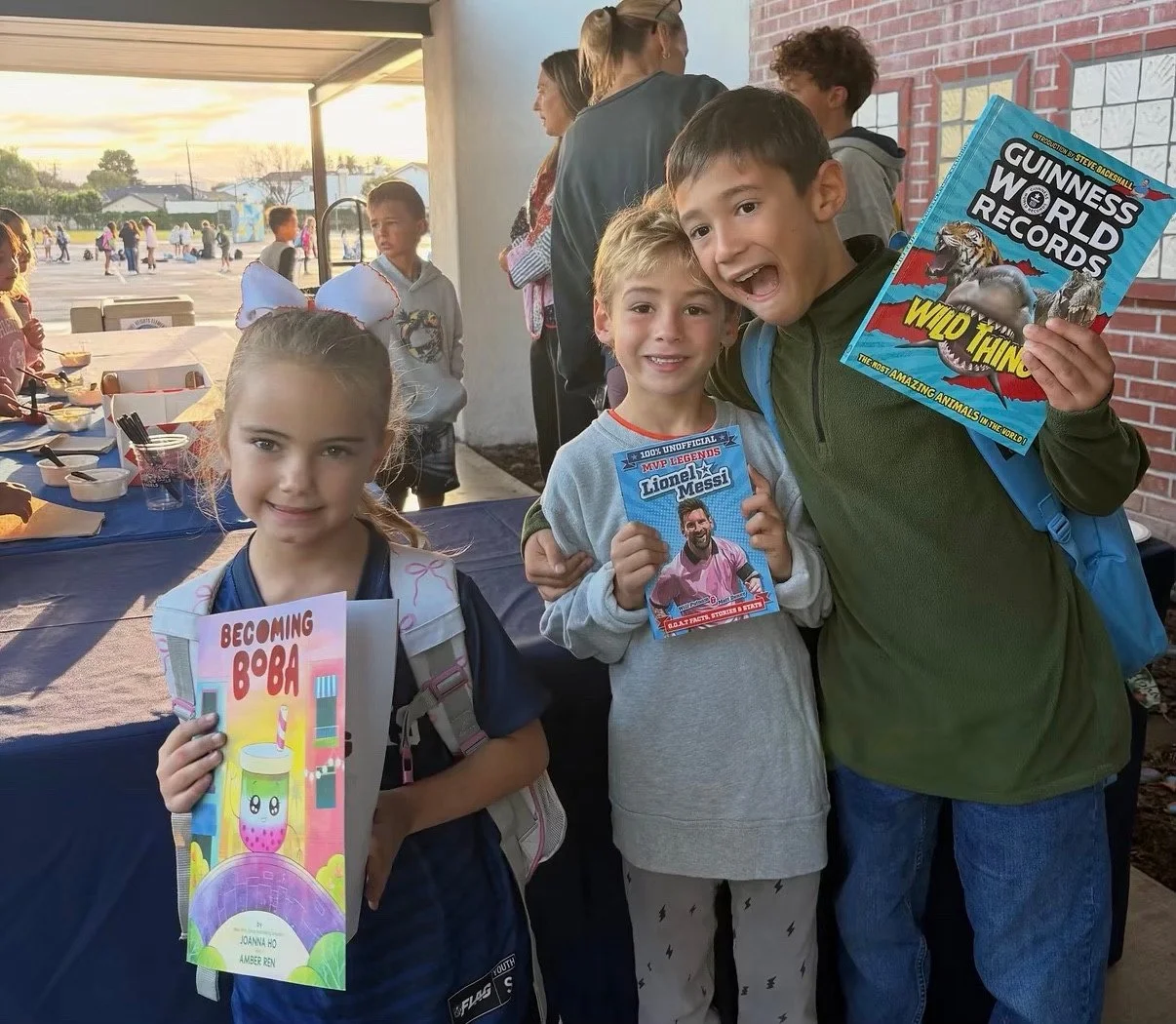 Three children with backpacks hold books outside a school building.