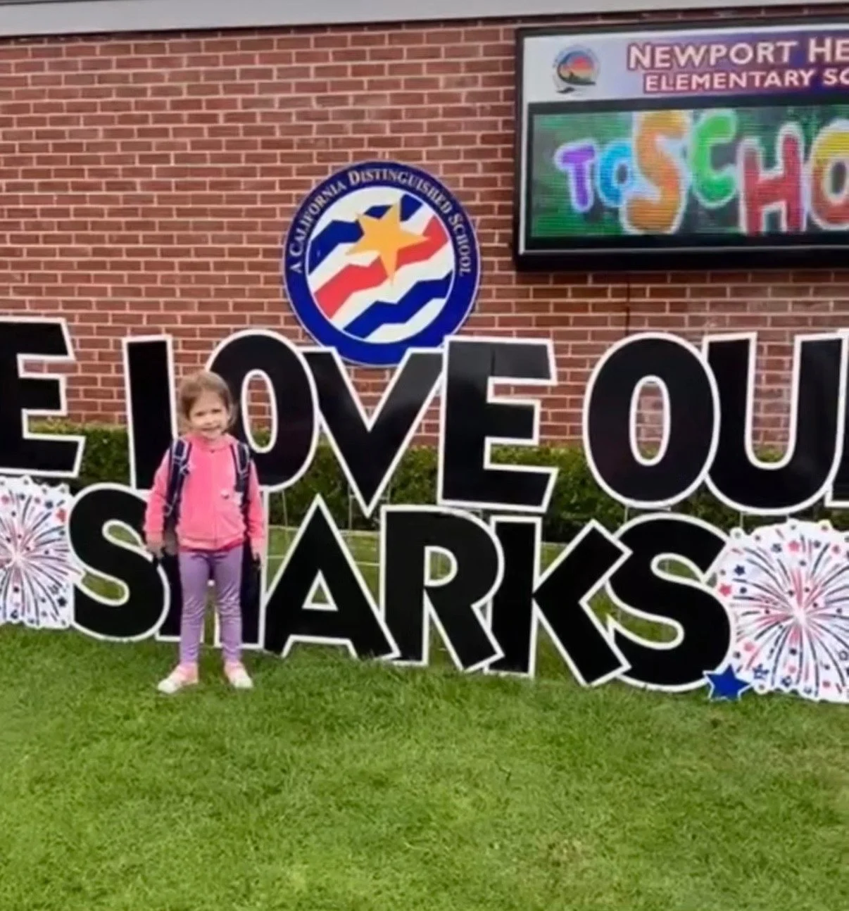 A child with a backpack stands beside a large Sharks welcome display outside the school.