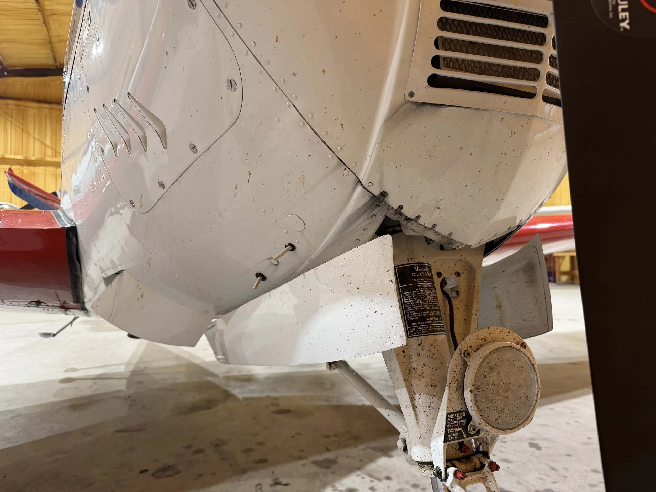 Close-up view of an aircraft's underbelly showing structural panels, rivets, and part of the landing gear assembly in a hangar.