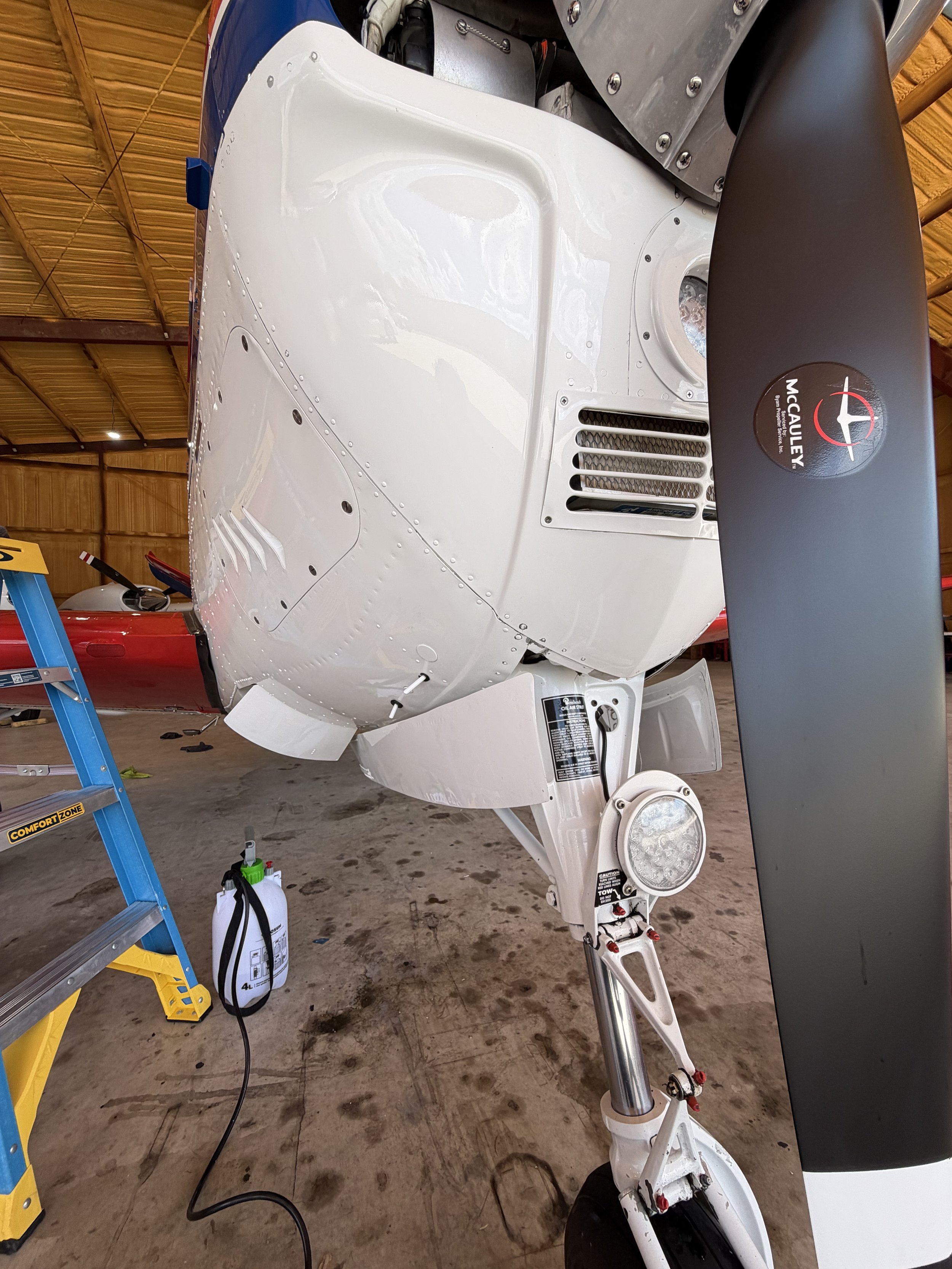 The image shows the side view of a partially assembled small aircraft or aircraft component in a hangar. The aircraft has a white fuselage with various bolts and openings, a black McAuley propeller attached, and some tools and supplies around it.