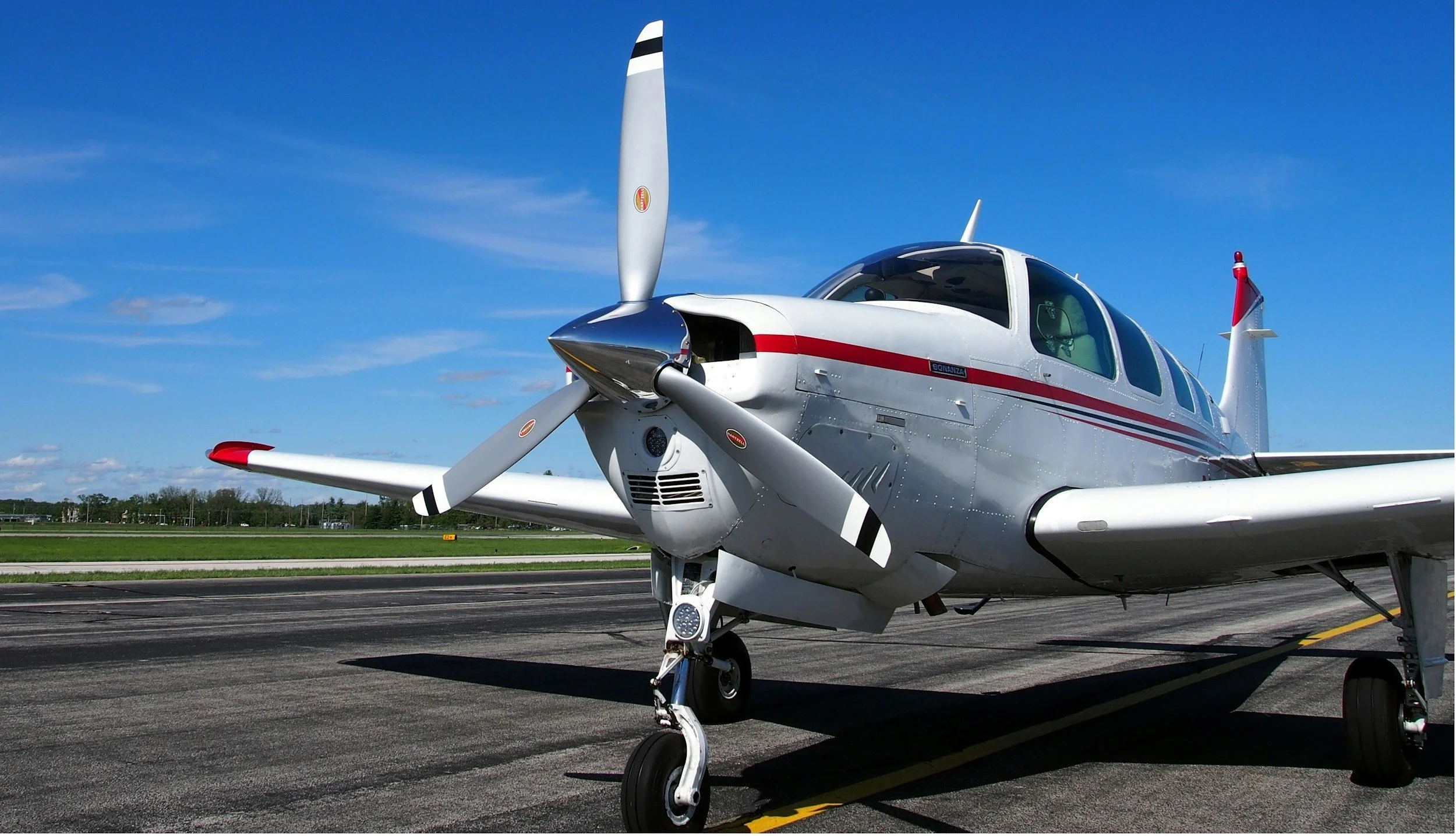 A white single-engine propeller airplane parked on an airport runway under a clear blue sky with a few clouds.