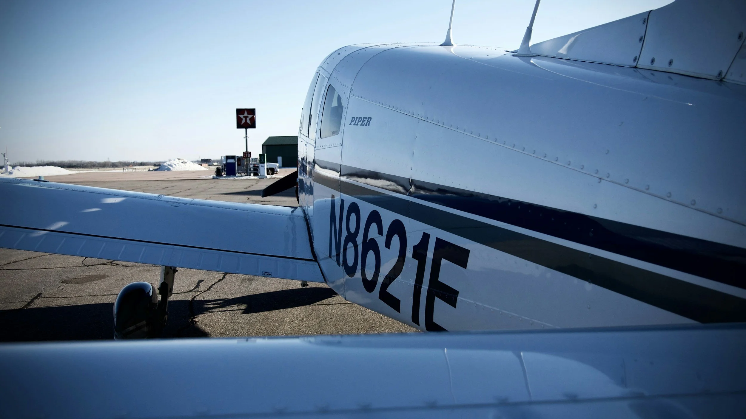 A small aircraft with the name 'Piper' on its side parked at a tarmac with fuel pumps, a green hangar, and snowy landscape in the background.
