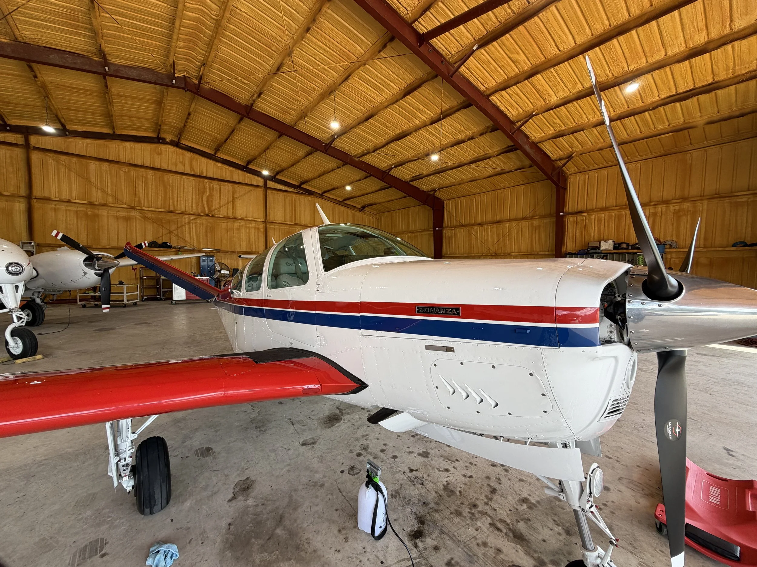 A small white aircraft with red and blue stripes inside a wooden airplane hangar.
