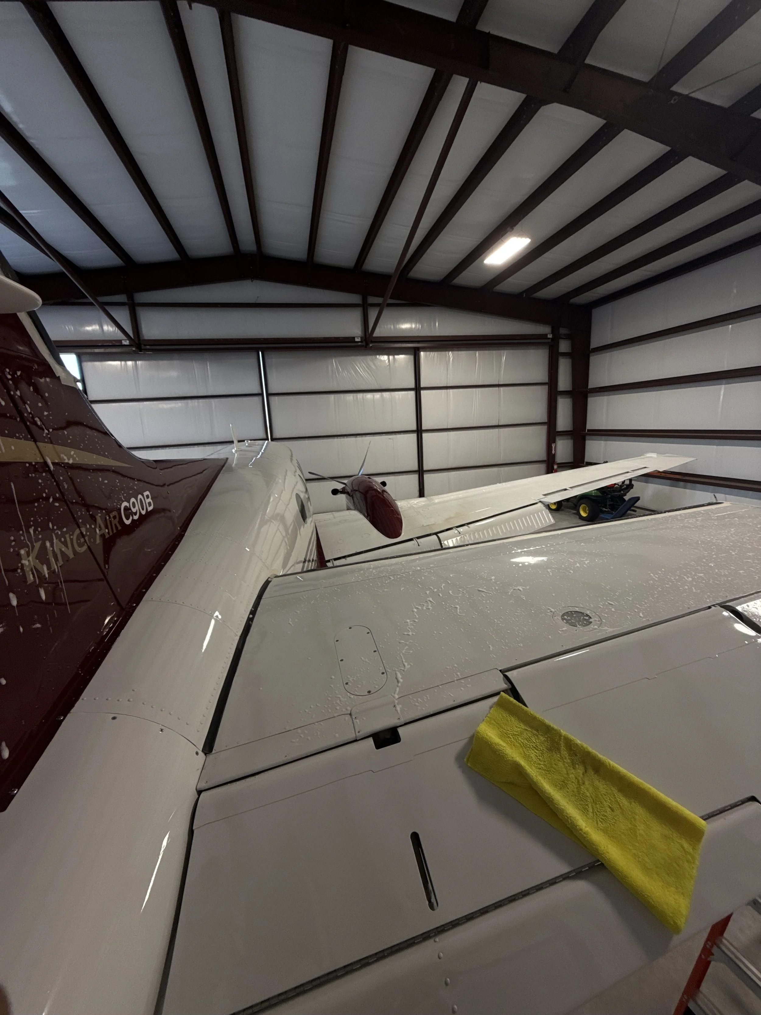 Inside a hangar with two small aircraft. The aircraft's wings are visible, and a yellow towel is draped over the wing in the foreground. The hangar has a metal roof and walls, and a light is turned on in the ceiling.