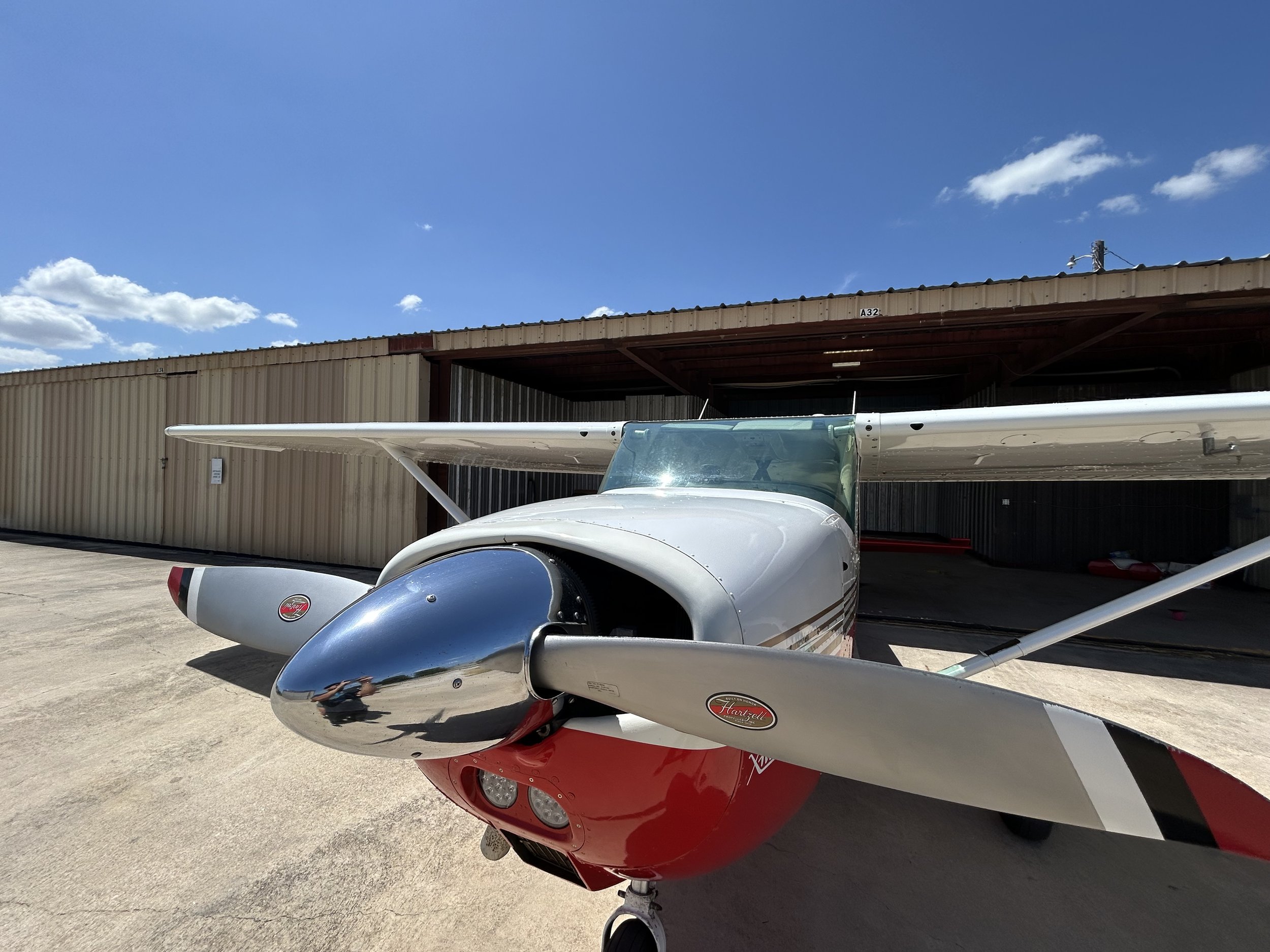 Small aircraft with white and red body parked in front of a beige hangar under a blue sky with some clouds.
