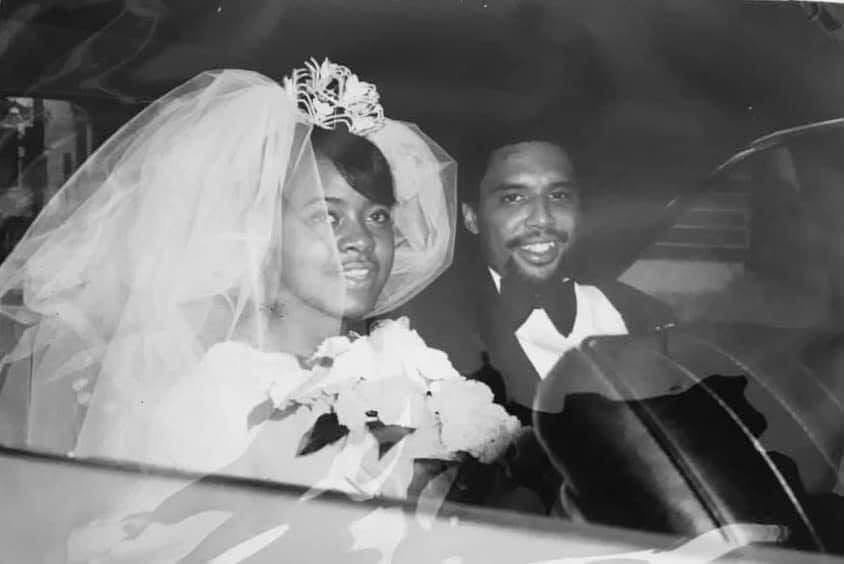Black-and-white photo of a bride and groom sitting in a car on their wedding day, with the bride wearing a veil and holding flowers, and the groom wearing a tuxedo, both smiling.