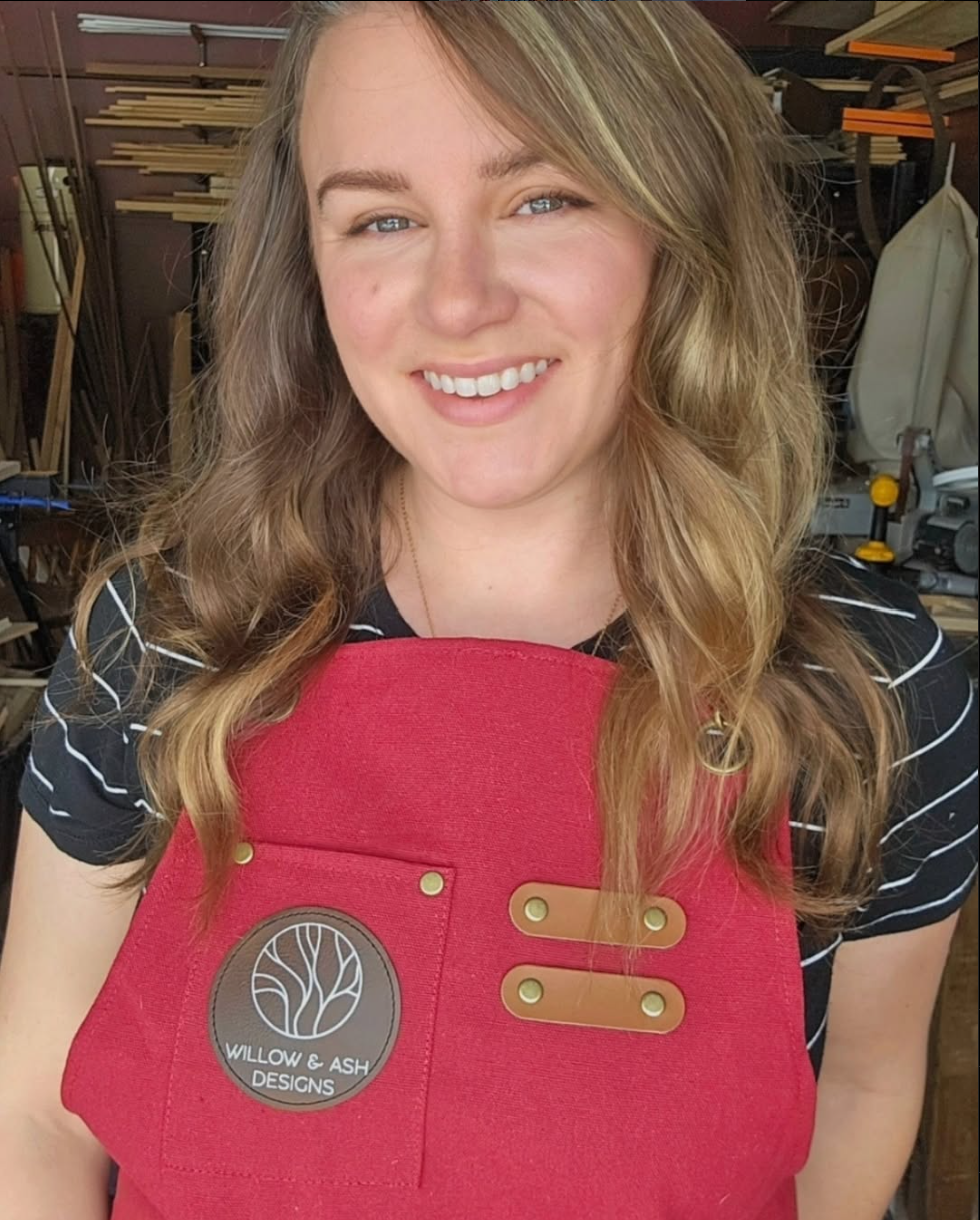 Ash, the wood artist, a woodworking workshop, wearing a red apron with a Willow & Ash Designs logo, and a black striped shirt, with woodworking tools visible in the background.