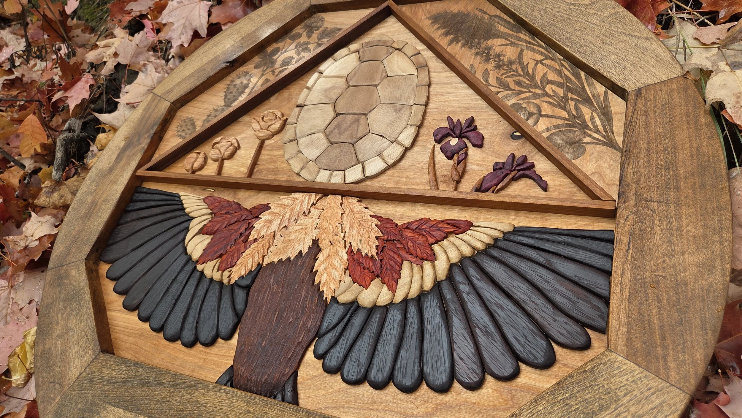 Wood inlay art of a bird with outstretched wings, surrounded by floral designs, on a wooden surface, with fallen autumn leaves around it.