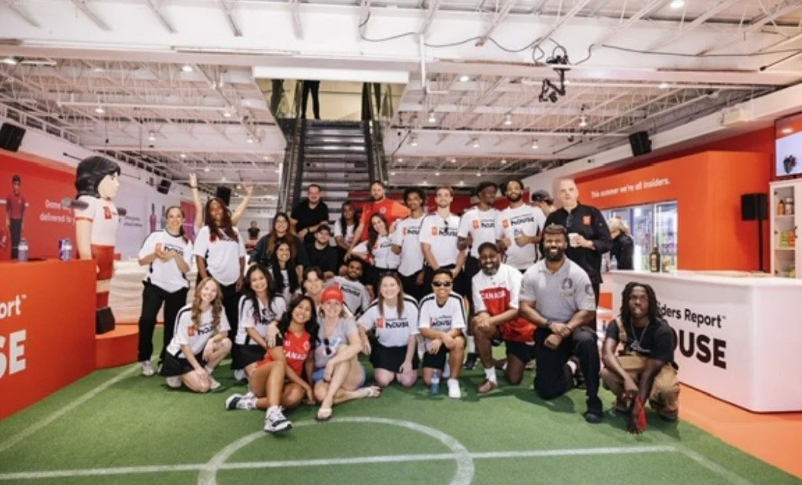 Large group posing inside a brightly lit sports store on an indoor turf field, surrounded by red and white displays.