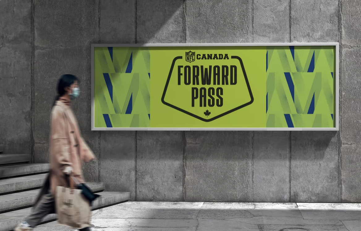 Masked pedestrian walking past an NFL Canada “Forward Pass” billboard in a concrete subway station