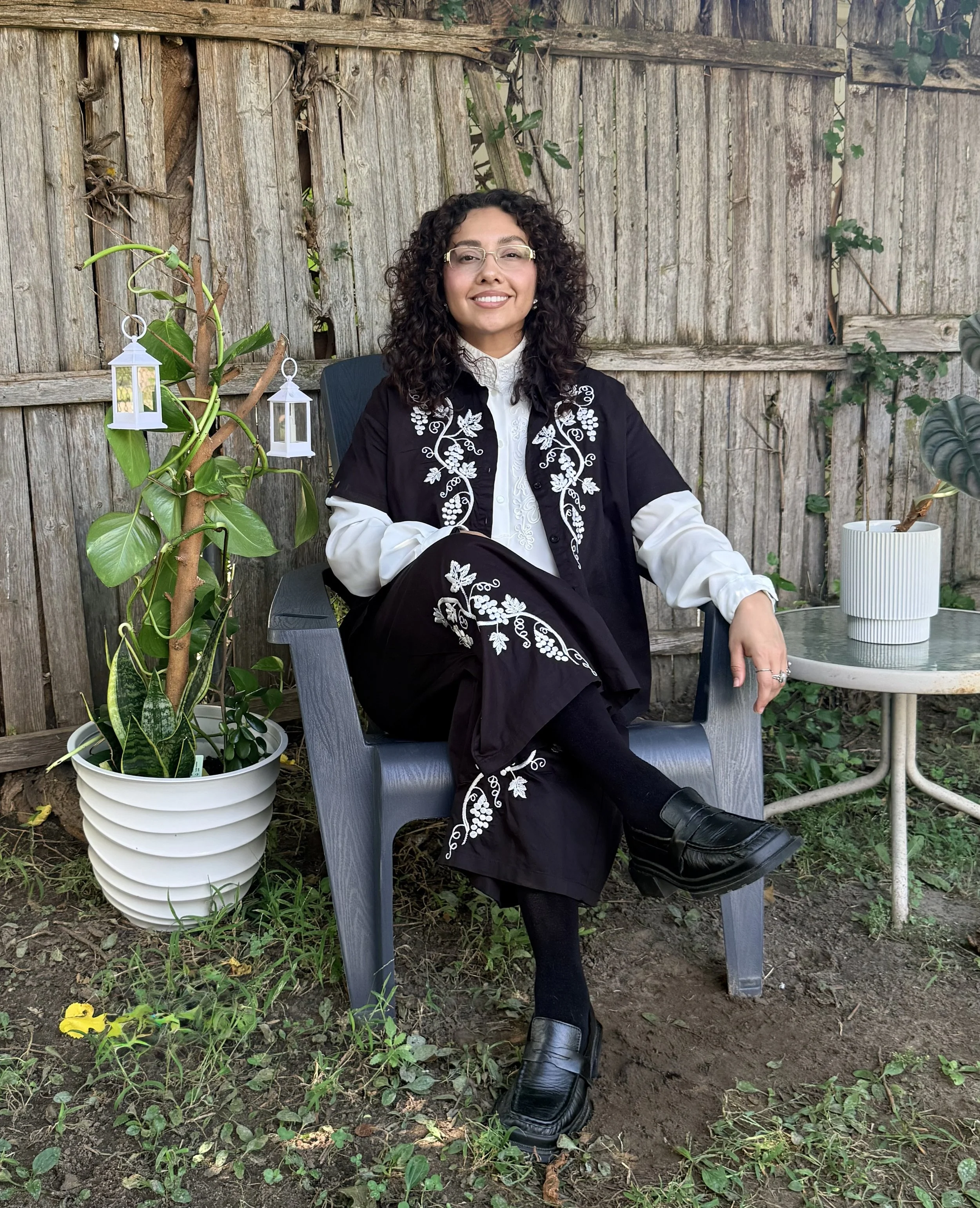 A woman with curly dark hair, glasses, and earrings sitting outdoors on a wooden chair in front of a weathered wooden fence. She is wearing a black and white embroidered outfit, black boots, and has one leg crossed over the other. There are potted plants nearby, including a large leafy plant with hanging lantern decorations, and yellow flowers on the ground.