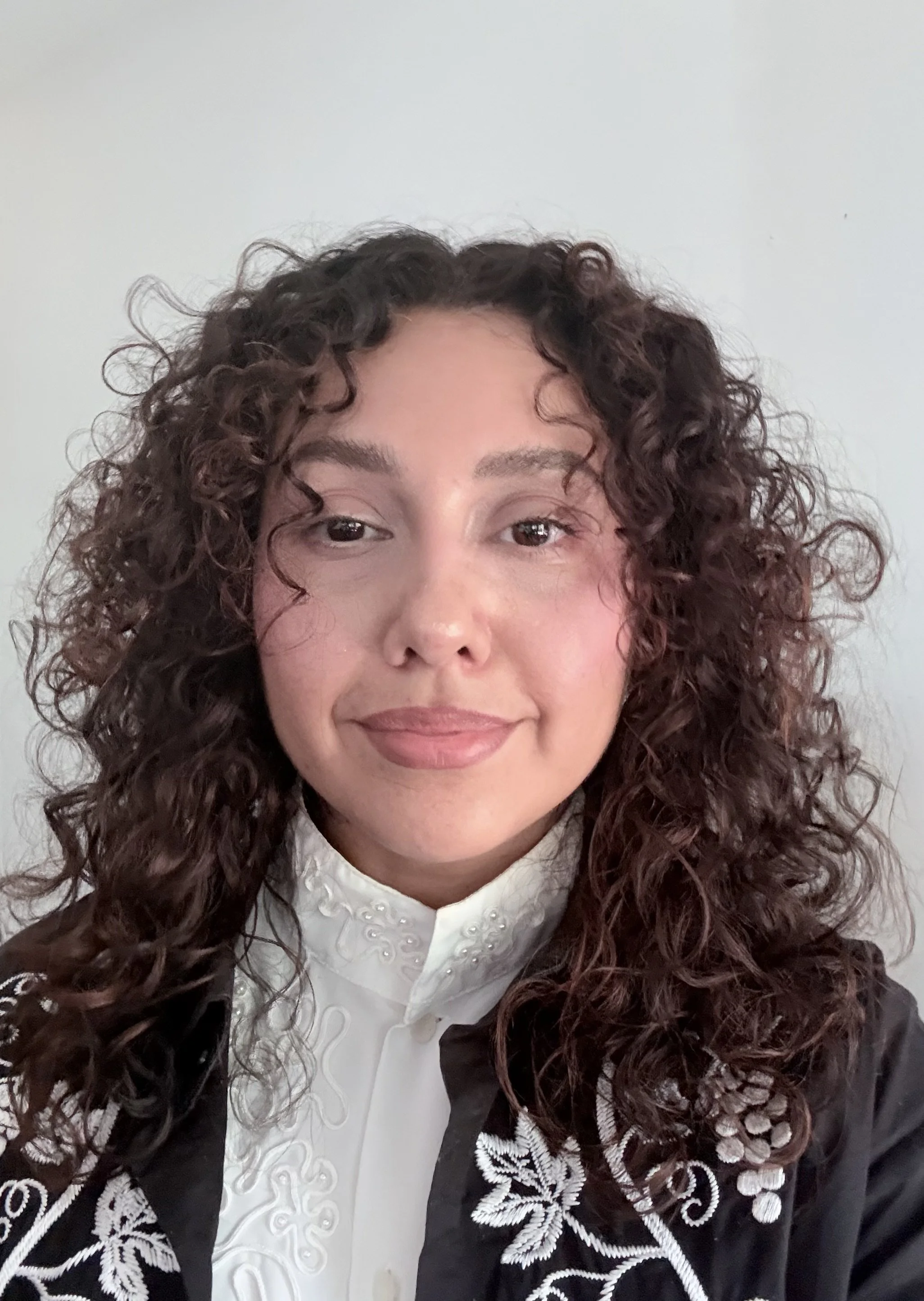 Close-up of a woman with curly dark hair wearing a white blouse with embroidery and a black jacket with white embroidery.
