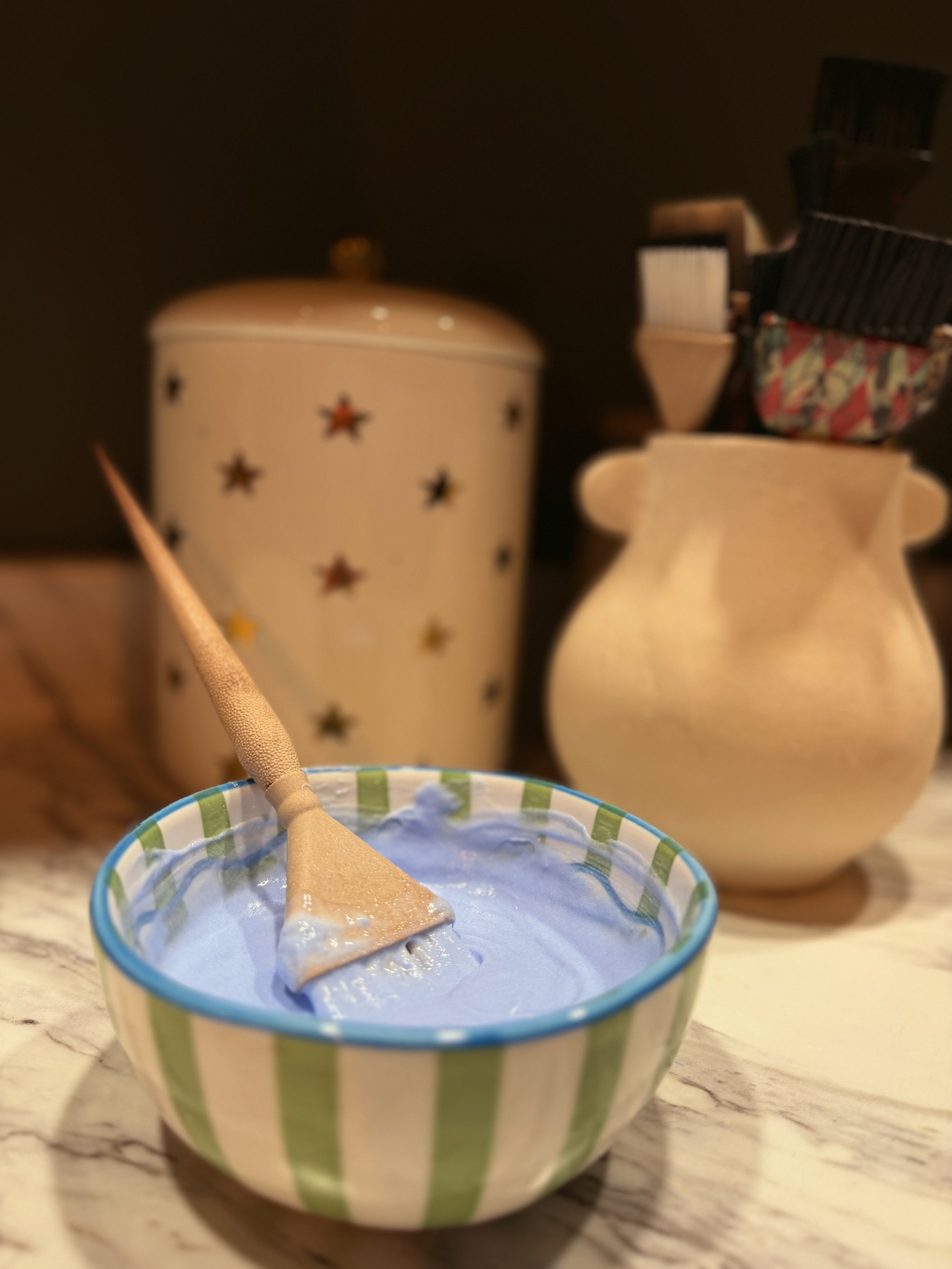 A cup with green and white stripes filled with a bleach  and a color brush resting inside, with blurred objects in the background.