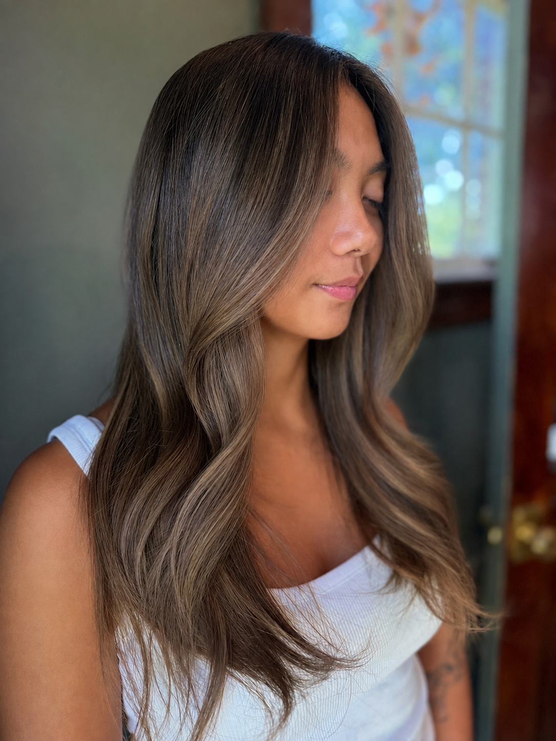 Woman with long, wavy hair, wearing a white top, standing indoors near a window with natural light.