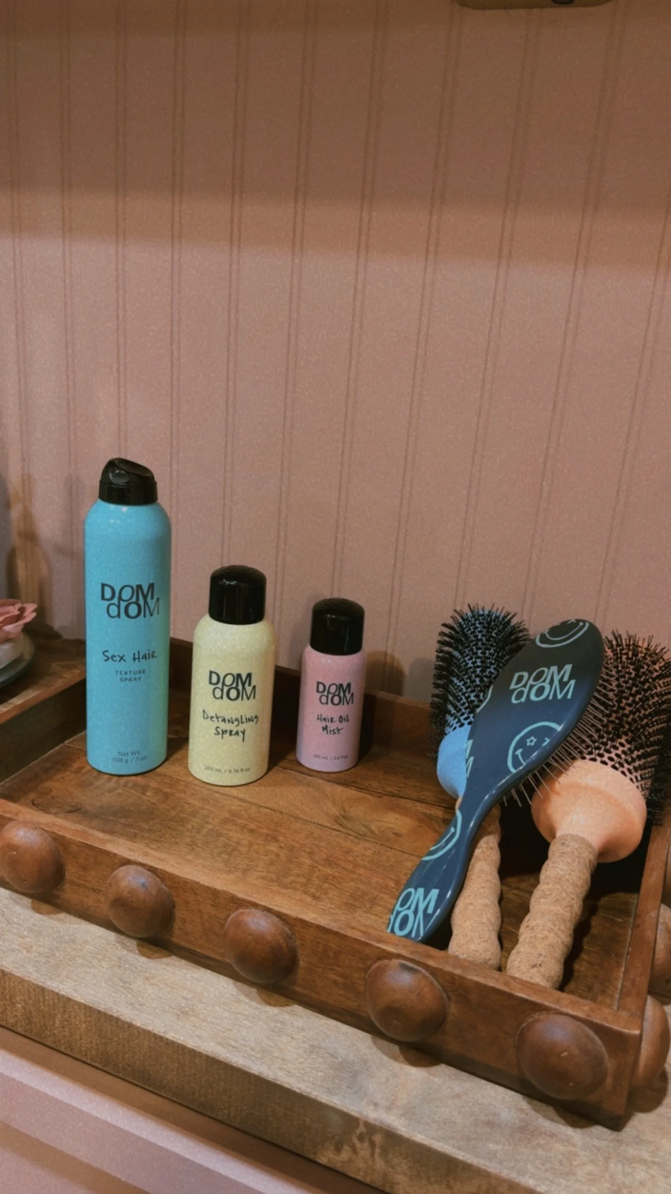 A wooden tray holding various hair care products including spray bottles labeled 'Sex Hair,' 'Detangling Spray,' and 'Hair Oil Mist,' along with hairbrushes and round objects.