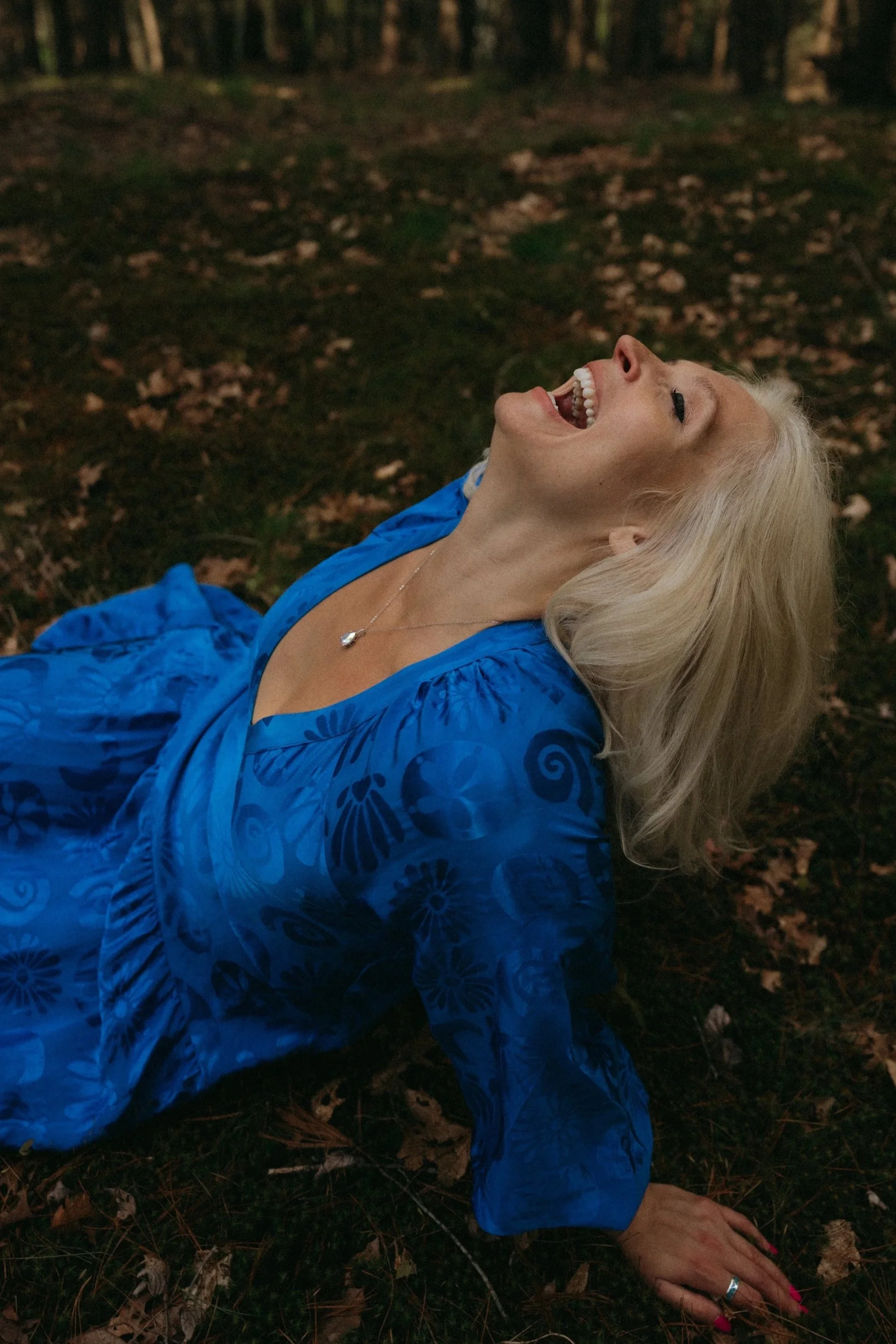 A woman with blonde hair, wearing a blue dress and a silver necklace, lying on the ground in a forest with leaves and moss, laughing with her head tilted back.