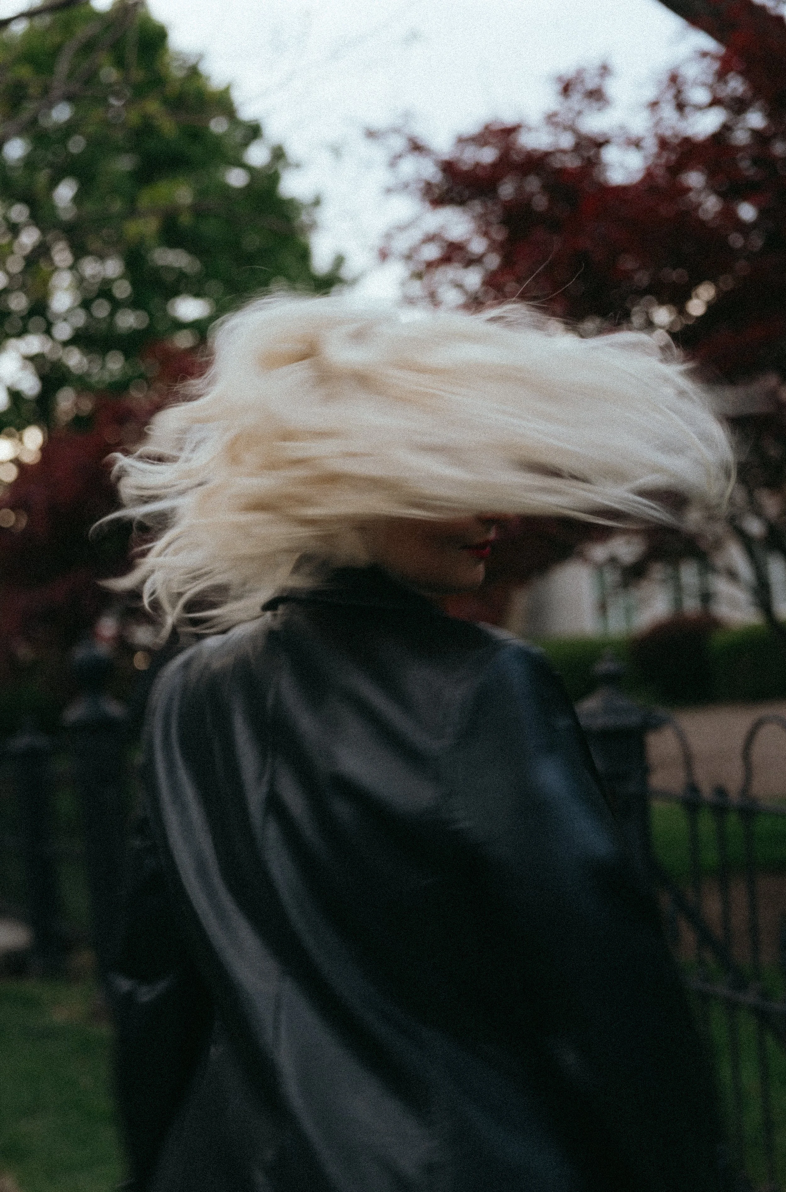 Person with platinum blonde hair, wearing a black leather jacket, standing outdoors in front of trees with green and red leaves, with a blurred background.