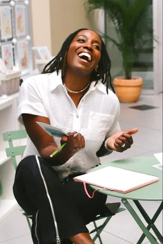 A woman with dreadlocks and a white shirt laughing openly, holding a card and a pen, sitting at a small round green table in an indoor setting with potted plants and shelves with magazines or books.