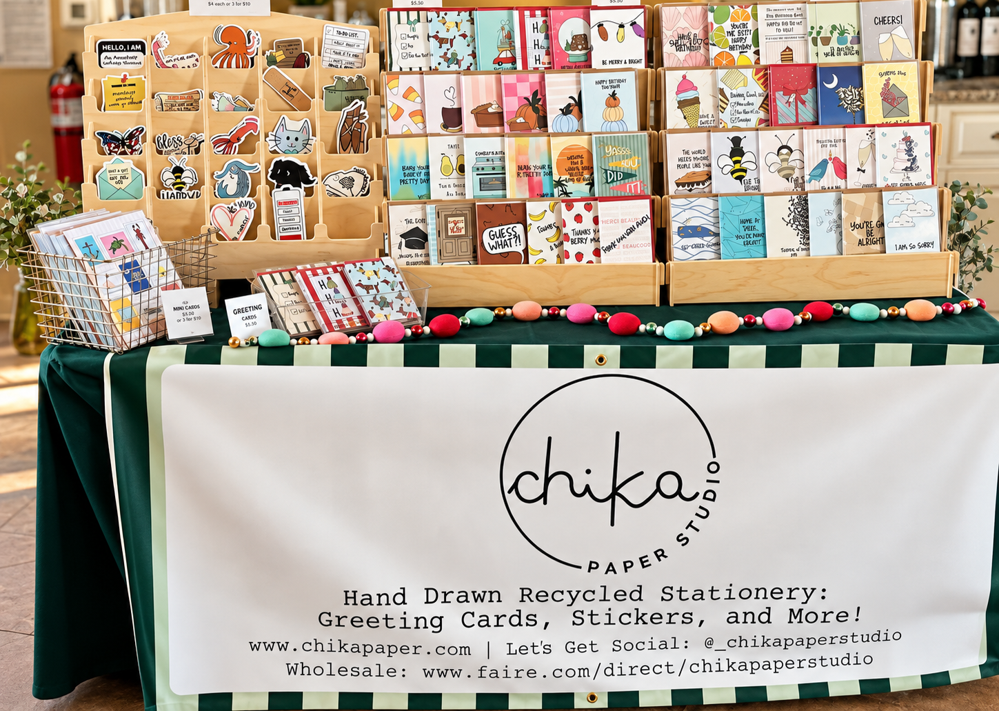 Display of hand-drawn, recycled stationery, including greeting cards and stickers, at a Chika Paper Studio booth with a green and white checkered tablecloth and a banner featuring the studio's logo and website.