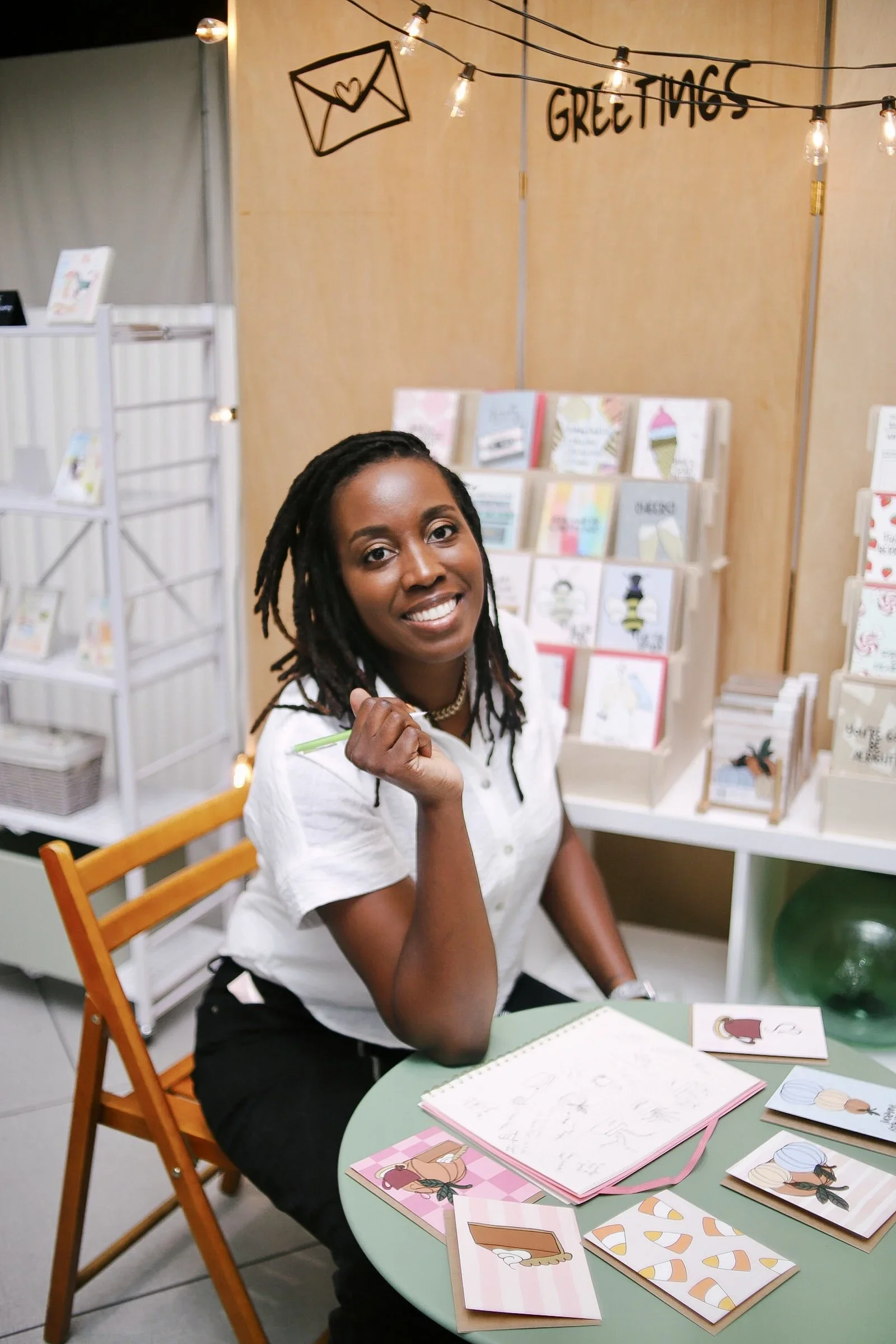 A woman with dark hair in long braids, wearing a white blouse, sitting at a green table with pink and white notebooks and greeting cards featuring illustrations of women with diverse hairstyles, while smiling and holding a green pen in her hand. Behind her, a display shelf with greeting cards and a wall with a sign reading 'GREETINGS' and an illustration of an envelope with a heart, is visible.