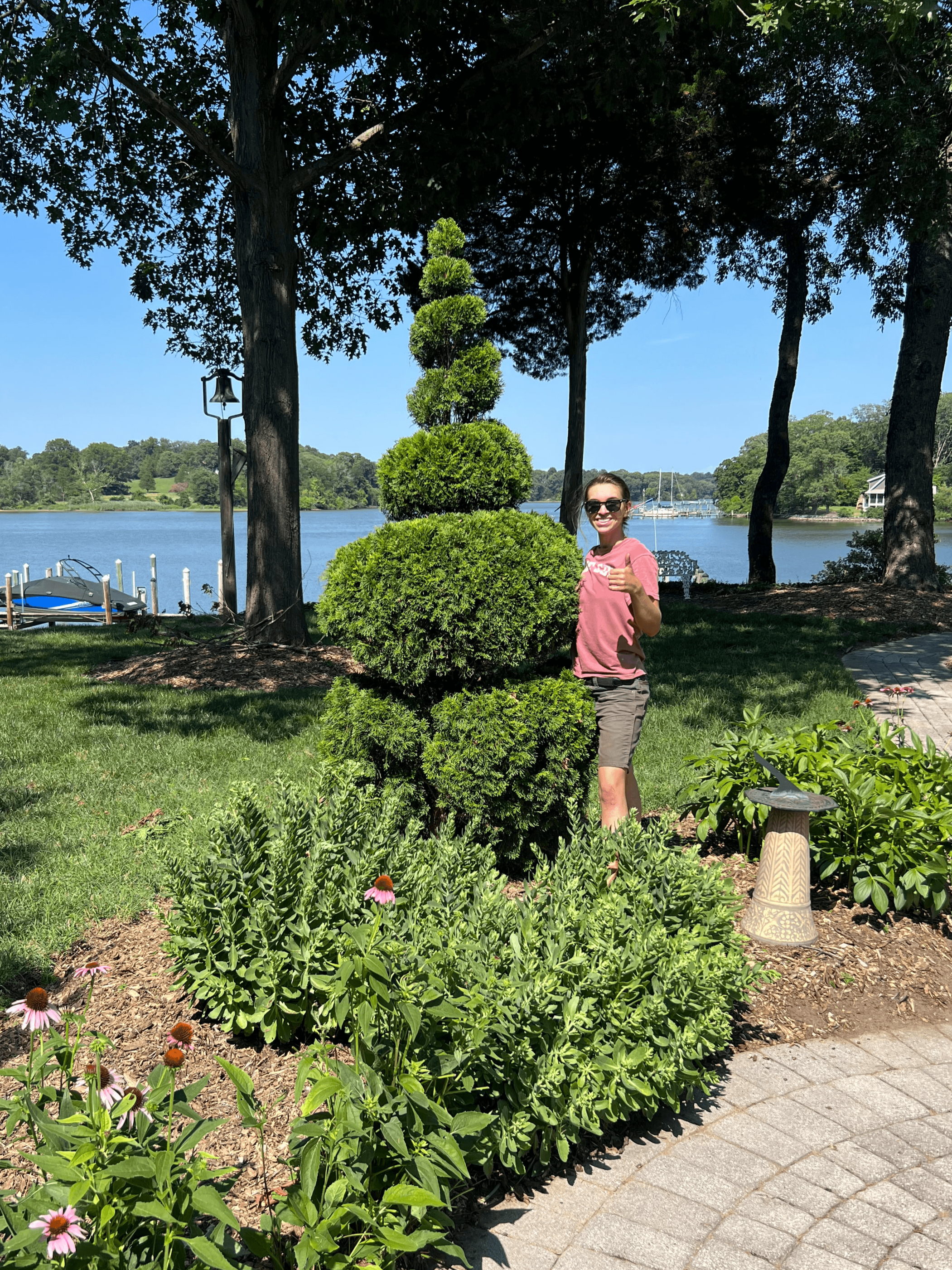 A woman with sunglasses giving a thumbs-up, standing in a garden near a waterfront with trees and boats in the background.