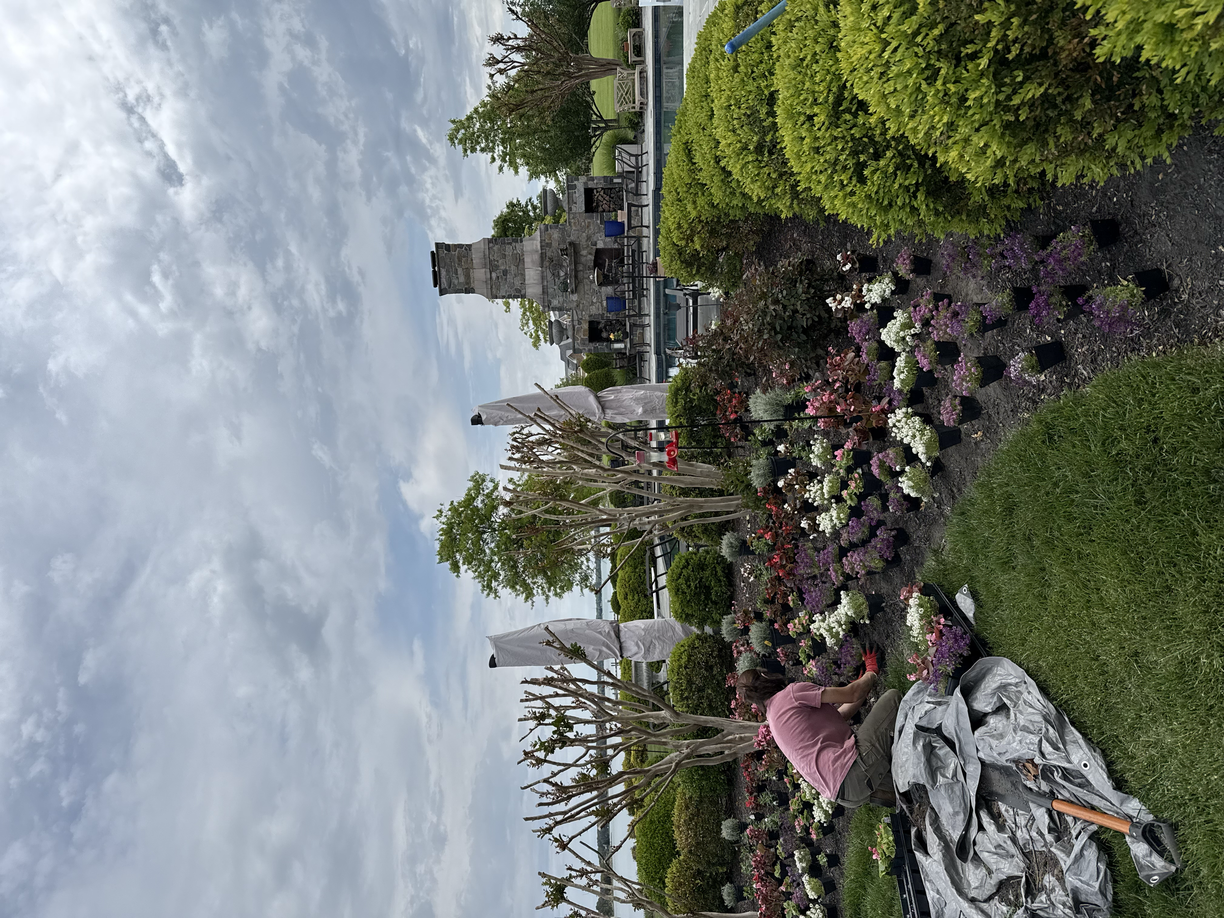 Person planting flowers in a garden with a variety of shrubs and trees, a stone fireplace, and outdoor furniture under a cloudy sky.