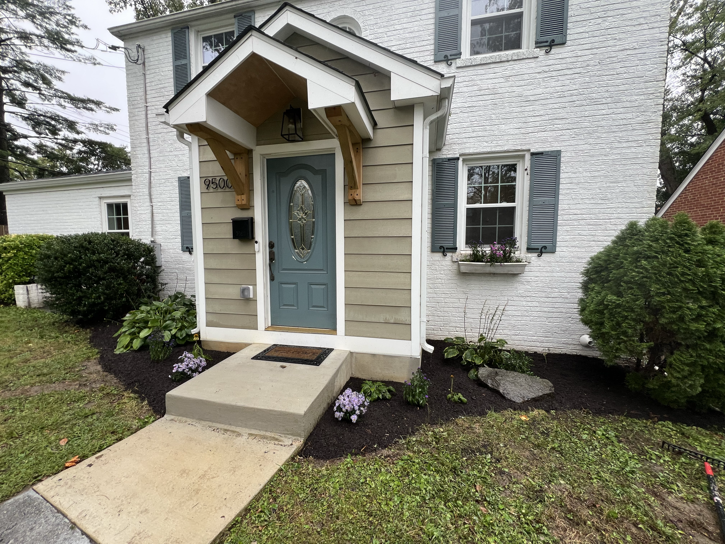 Front entrance of a house with a blue door, beige siding, white brick wall, black shutters, a small porch with a wooden awning supported by braces, a lantern light, and a black mailbox. There are flowering plants and bushes in the garden.