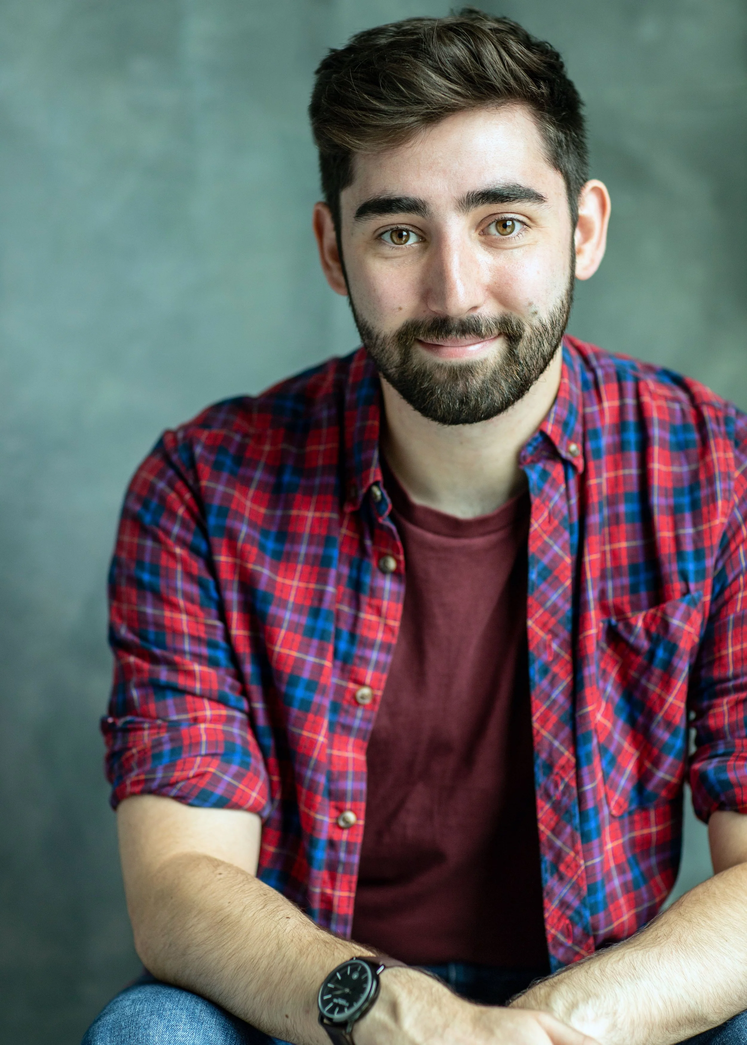 A young man with brown hair and a beard sitting against a gray background, wearing a red plaid shirt over a maroon t-shirt, with a black watch on his left wrist, smiling at the camera.
