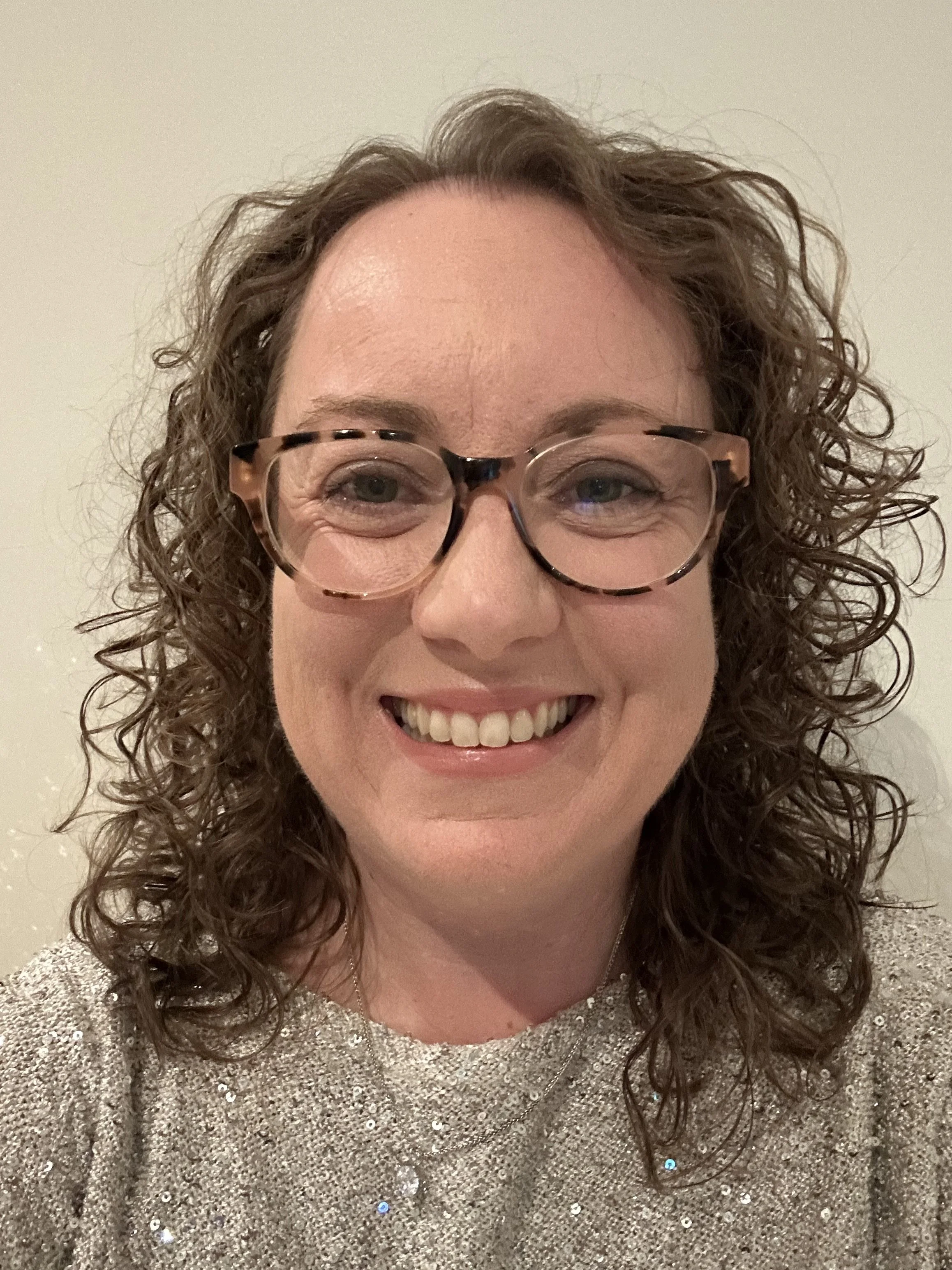 Close-up of a smiling woman with curly hair wearing tortoiseshell glasses and a glittering silver top.