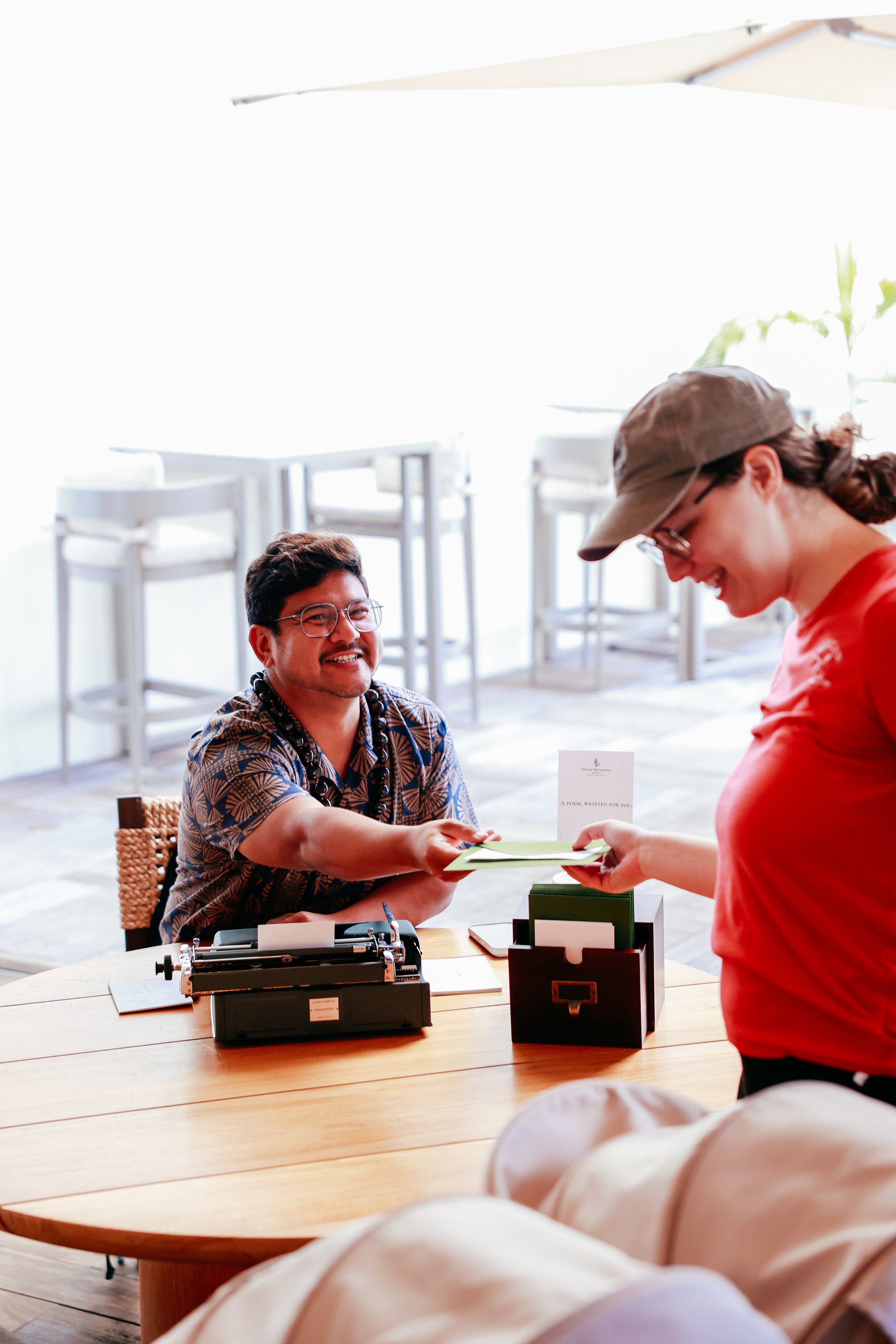 A man with glasses and a Hawaiian shirt sits at a wooden table, smiling and shaking hands with a woman wearing a hat and red shirt in a bright, modern indoor space. There is a typewriter and a mailbox on the table.