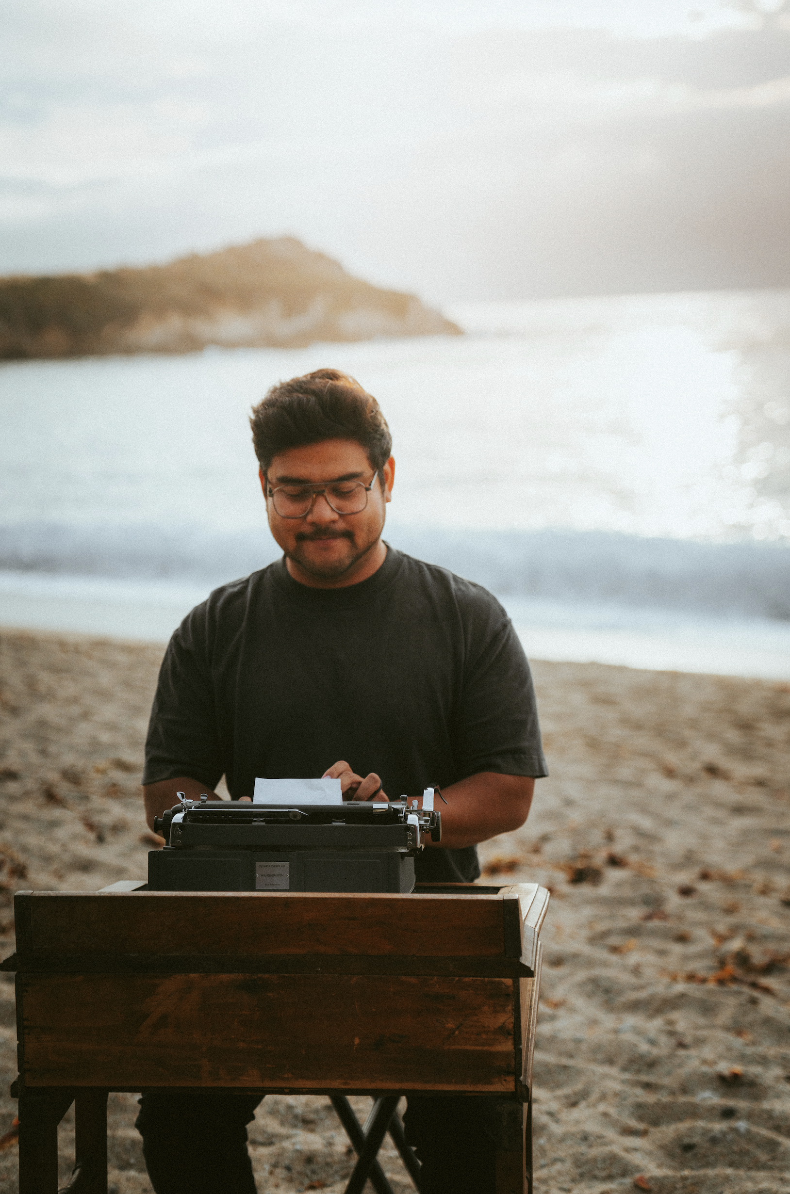 A man with glasses and dark hair, wearing a black shirt, standing on a sandy beach, operating a vintage typewriter on a wooden desk, with the ocean and a distant rocky coastline in the background during sunset.