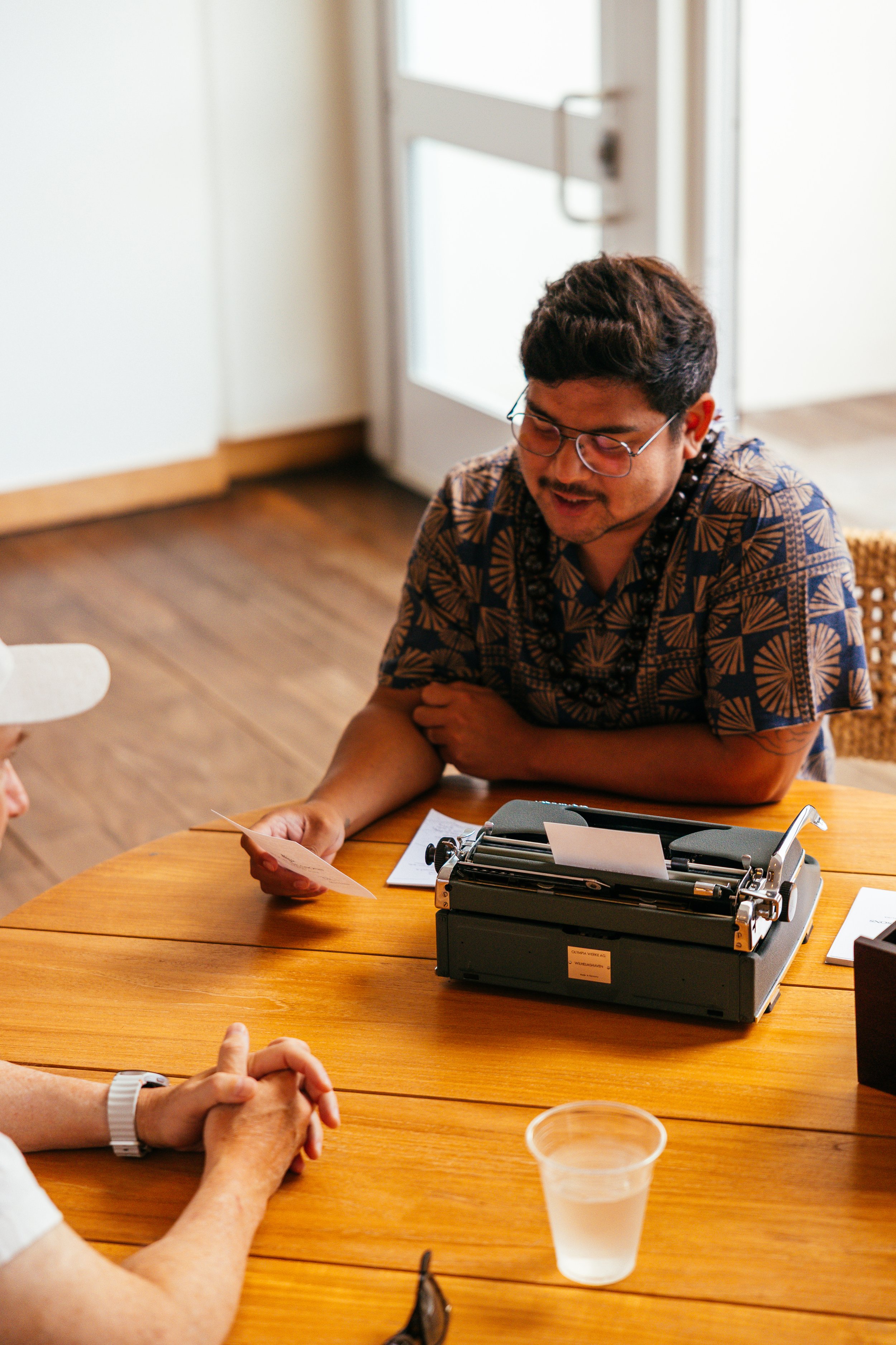 A man with glasses sitting at a wooden table, looking at a piece of paper. There is a typewriter on the table in front of him, and other people are partially visible, including someone with clasped hands and a white watch and another person with a translucent cup filled with water.
