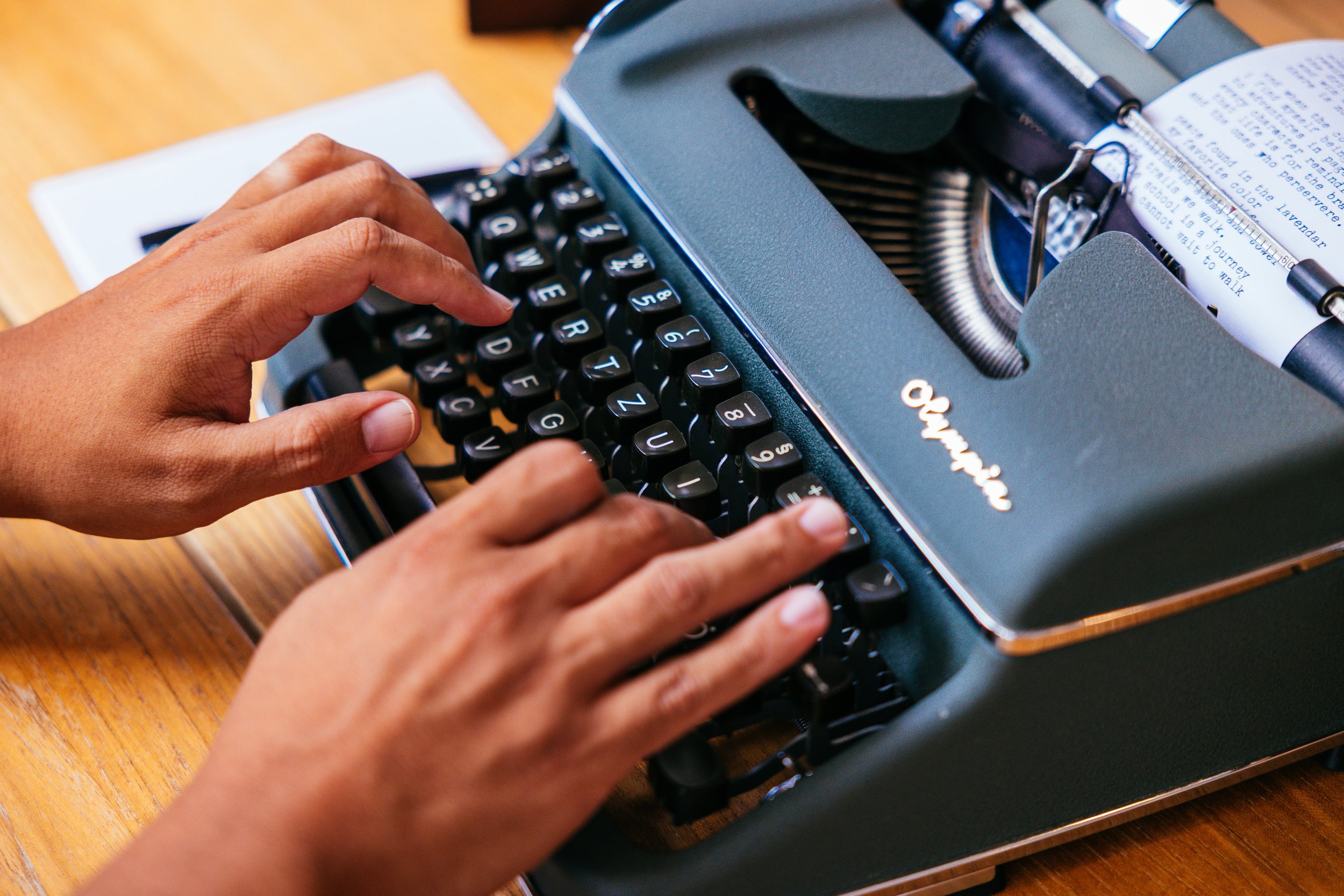 Close-up of a person's hands typing on a vintage black typewriter on a wooden desk with a book or paper in the background.